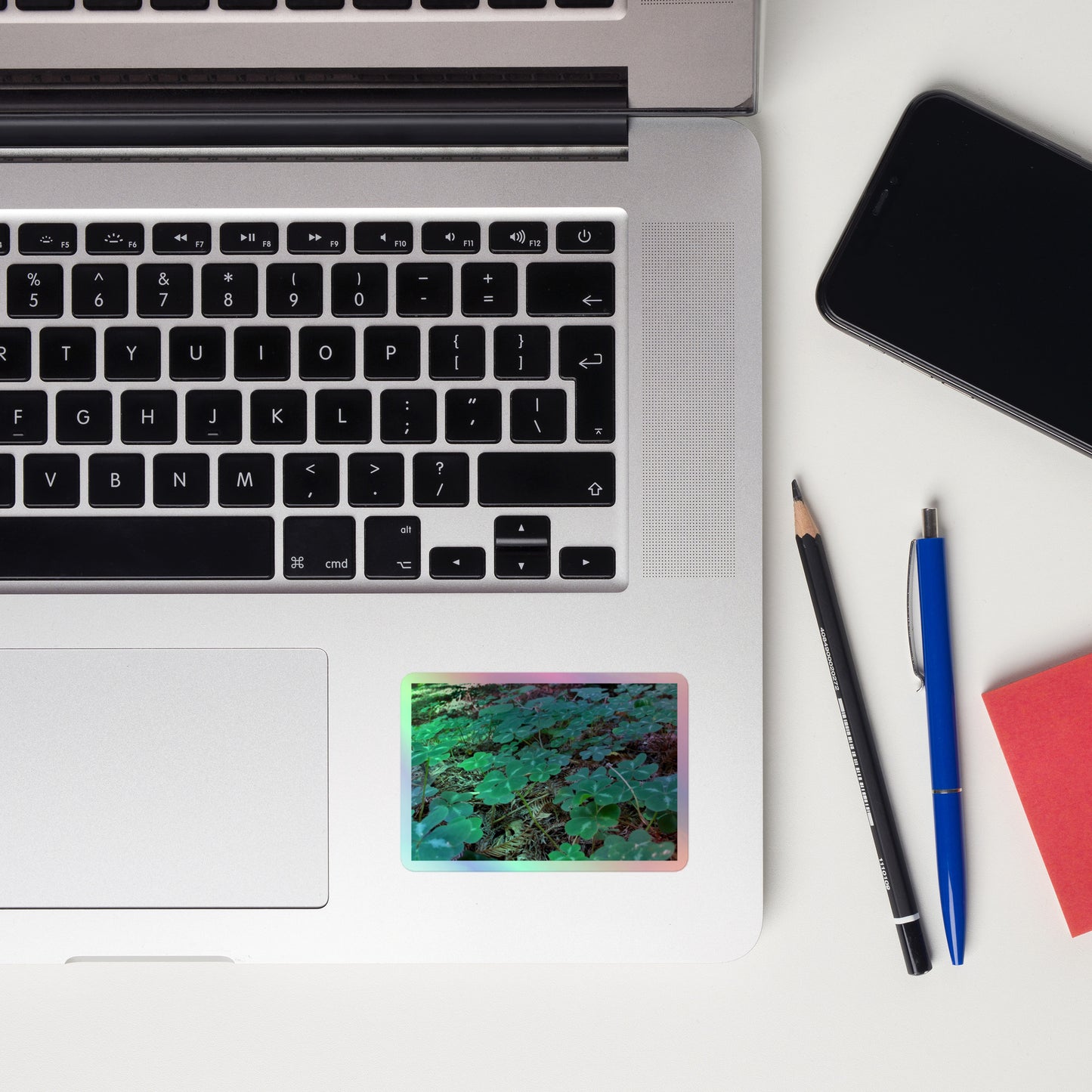 Lifestyle photo of a rectangular holographic sticker with an image of vivid green clover among redwood needles, placed on a laptop to the right of the mouse pad.