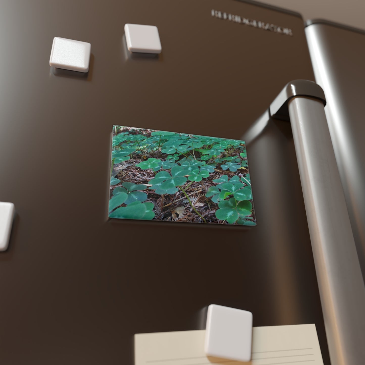 Lifestyle photo of a rectangular button magnet with an image of a vivid green field of clover amidst redwood needles, placed on a fridge.