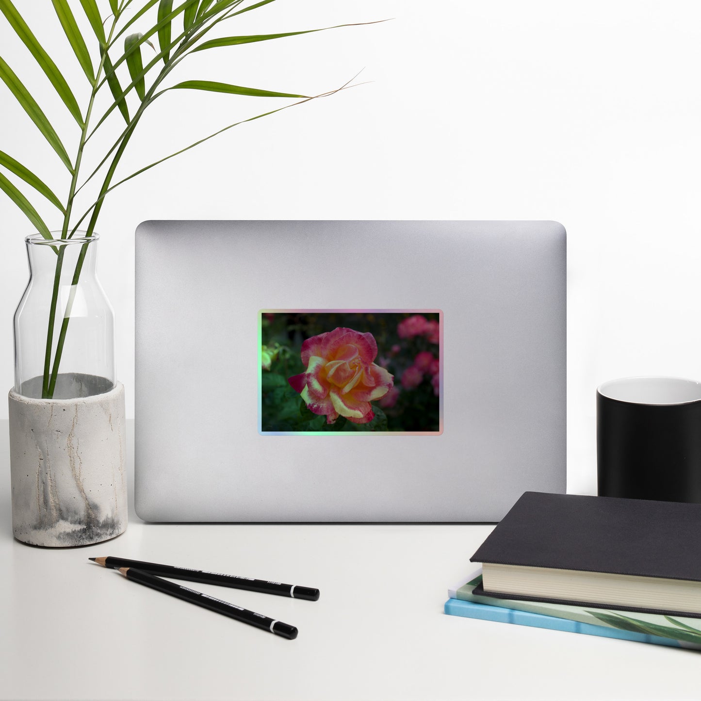 Lifestyle photo of a rectangular holographic sticker with a close-up image of a close-up image of pink and yellow rose bloom in a garden, placed on a laptop with desk supplies nearby.