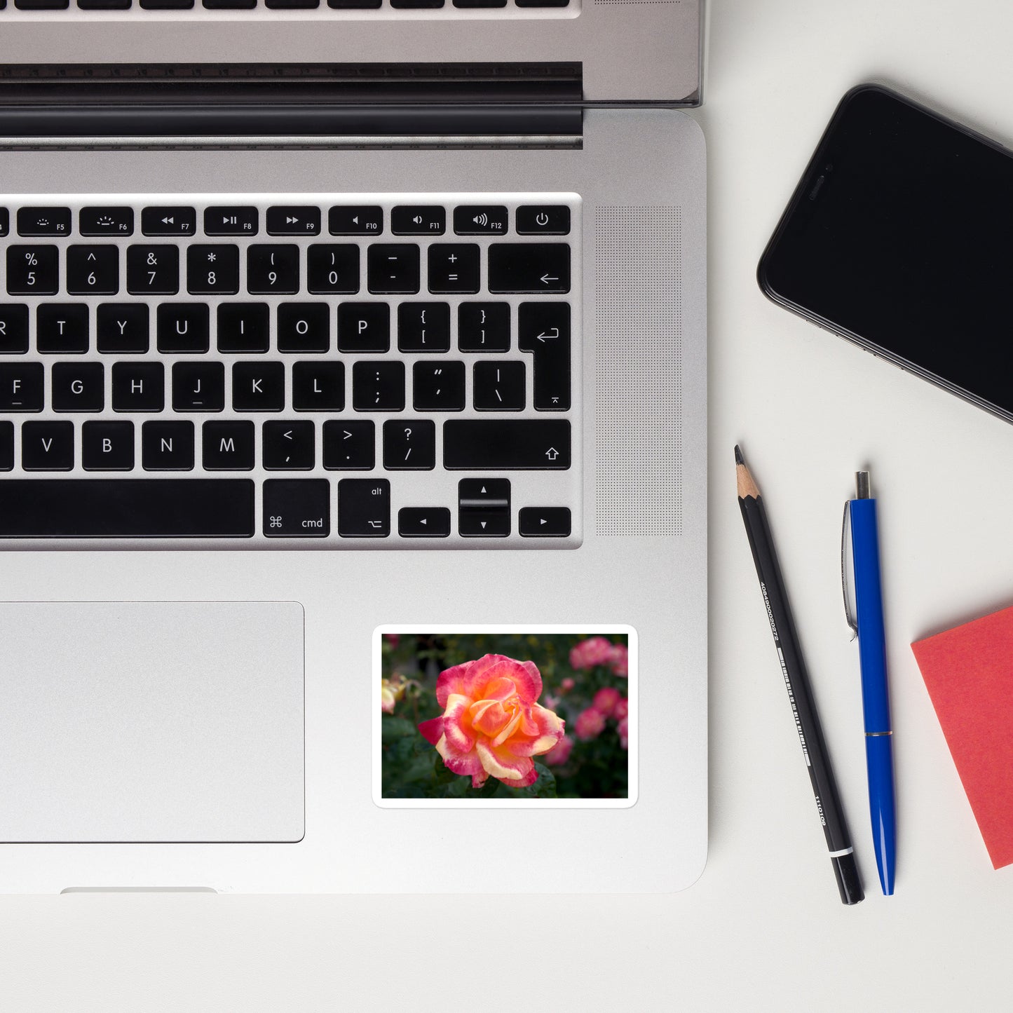 Lifestyle photo of a rectangular vinyl sticker with an image of a pink and yellow rose bloom with dark green leaves, placed on a laptop to the right of the mouse pad.