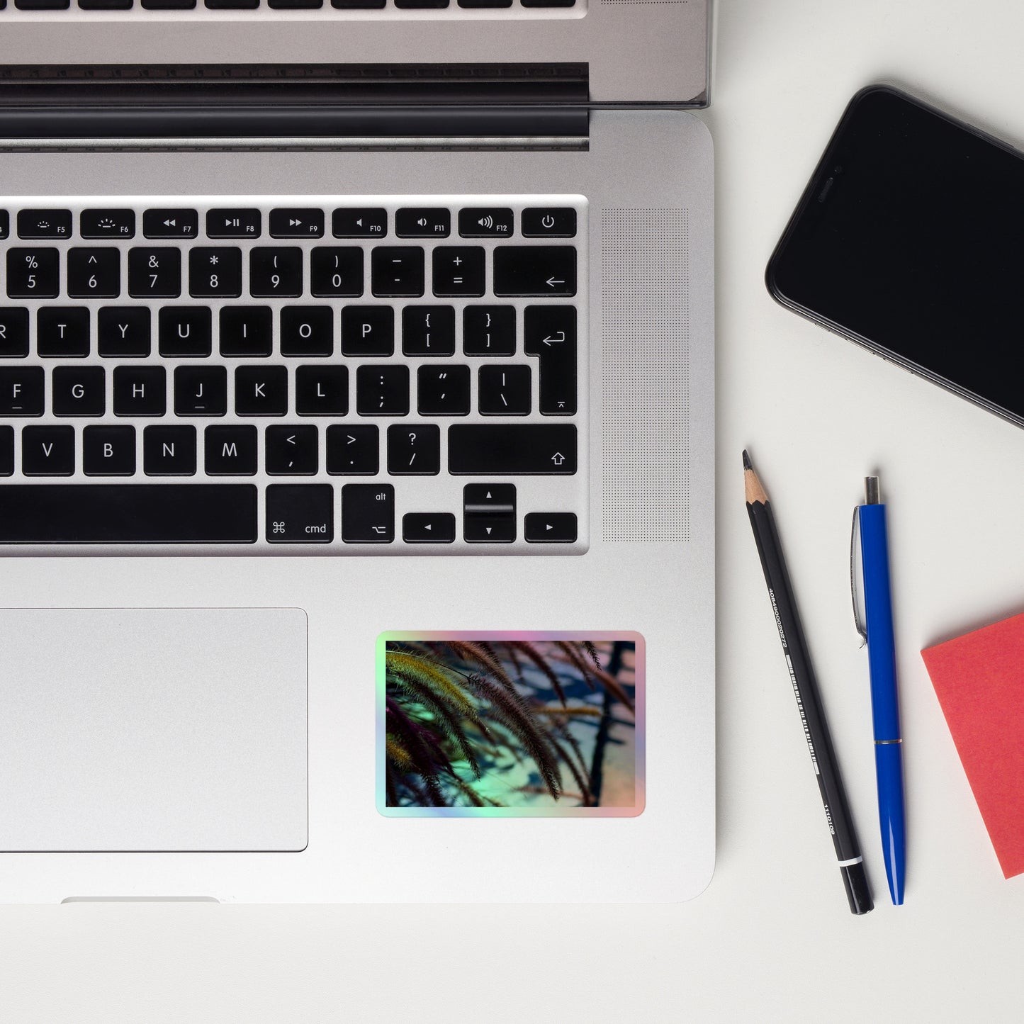 Lifestyle photo of a rectangular holographic sticker with an image ofgolden purple foxtails breeze, placed on a laptop to the right of the mouse pad.