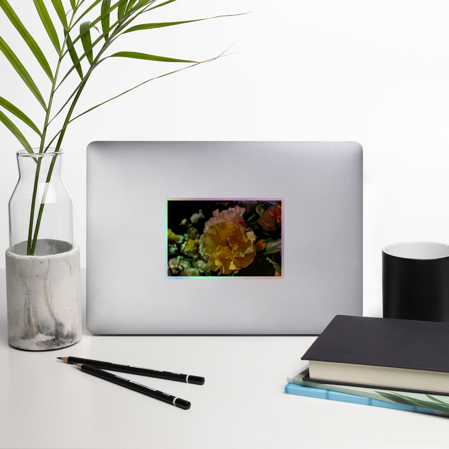 Lifestyle photo of a rectangular holographic sticker with an image of pale yellow and pink rose blooms in a garden, placed on a laptop with desk supplies nearby.