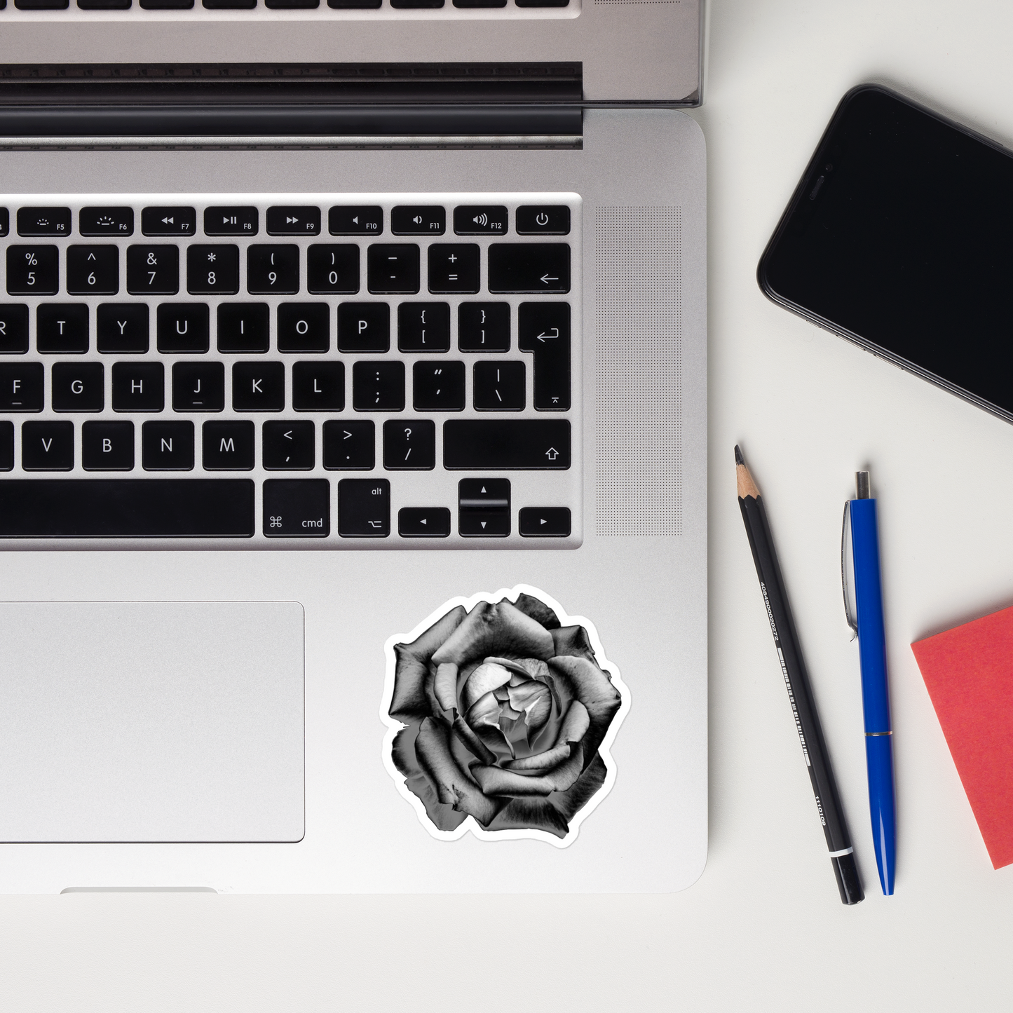 Lifestyle photo of a kiss-cut vinyl sticker with an image of a grey charcoal drawing style rose, placed on a laptop to the right of the mouse pad.