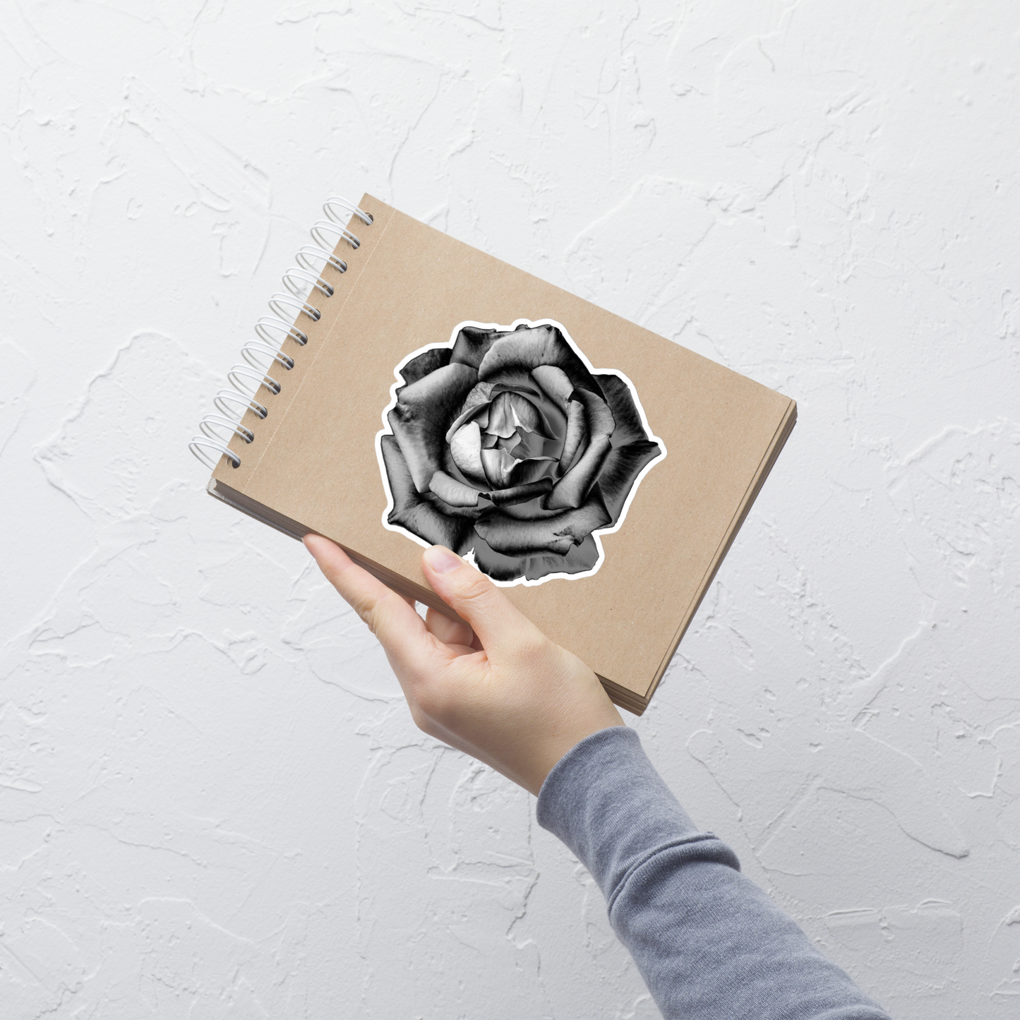 Lifestyle photo of a kiss-cut vinyl sticker with an image of a grey charcoal drawing style rose, placed on a small notepad.