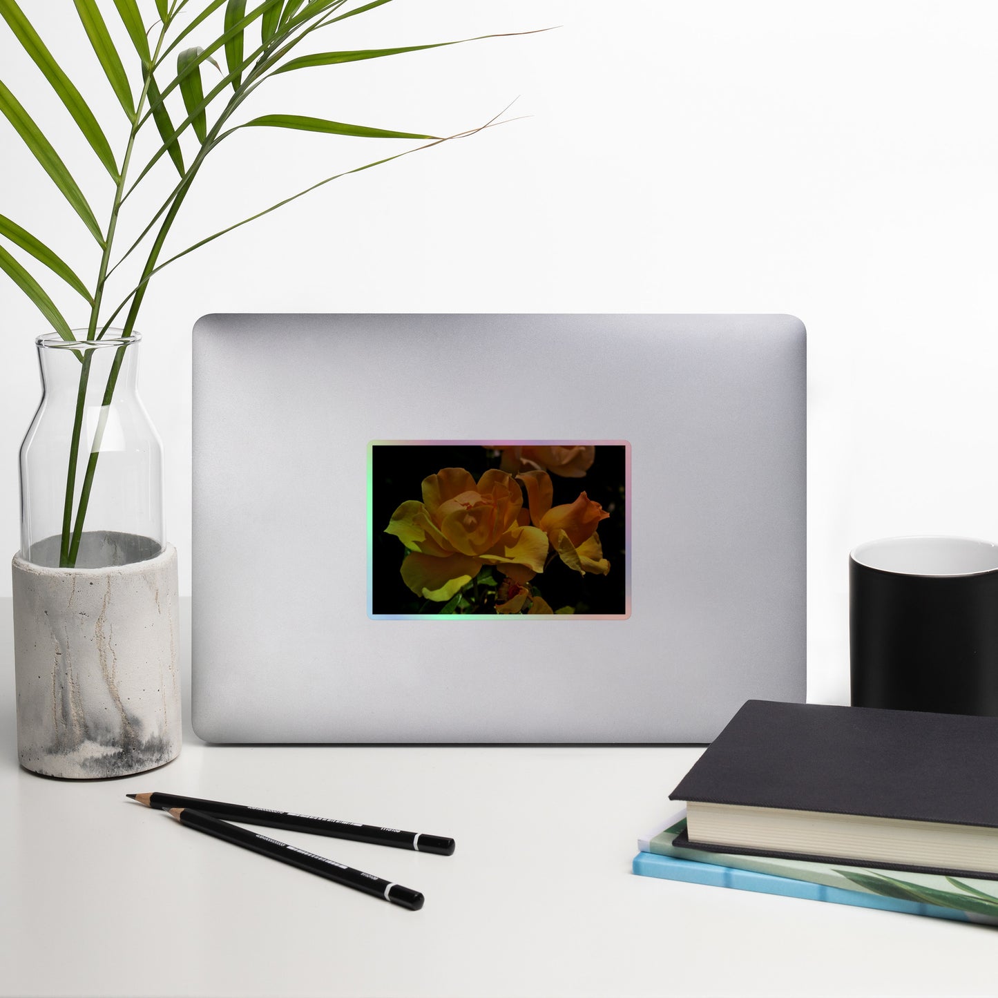 Lifestyle photo of a rectangular holographic sticker with a high-contrast image of vivid yellow roses against a dark background, placed on a laptop with desk supplies nearby.
