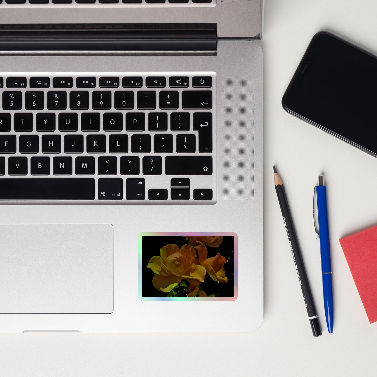Lifestyle photo of a rectangular holographic sticker with a high-contrast image of vivid yellow roses against a dark background, placed on a laptop to the right of the mouse pad.