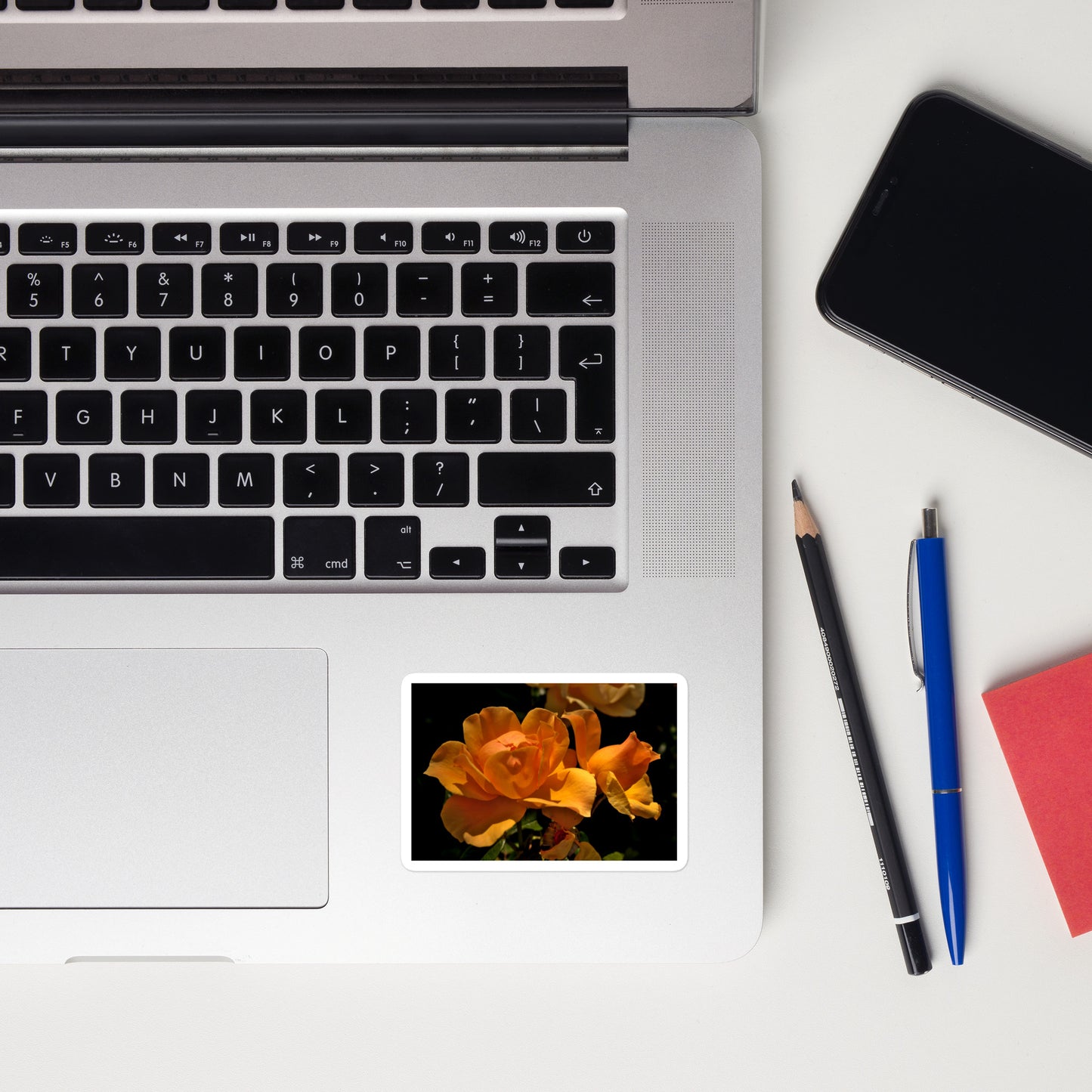 Lifestyle photo of a rectangular vinyl sticker with a high-contrast image of vivid orange roses against a dark background, placed on a laptop to the right of the mouse pad.