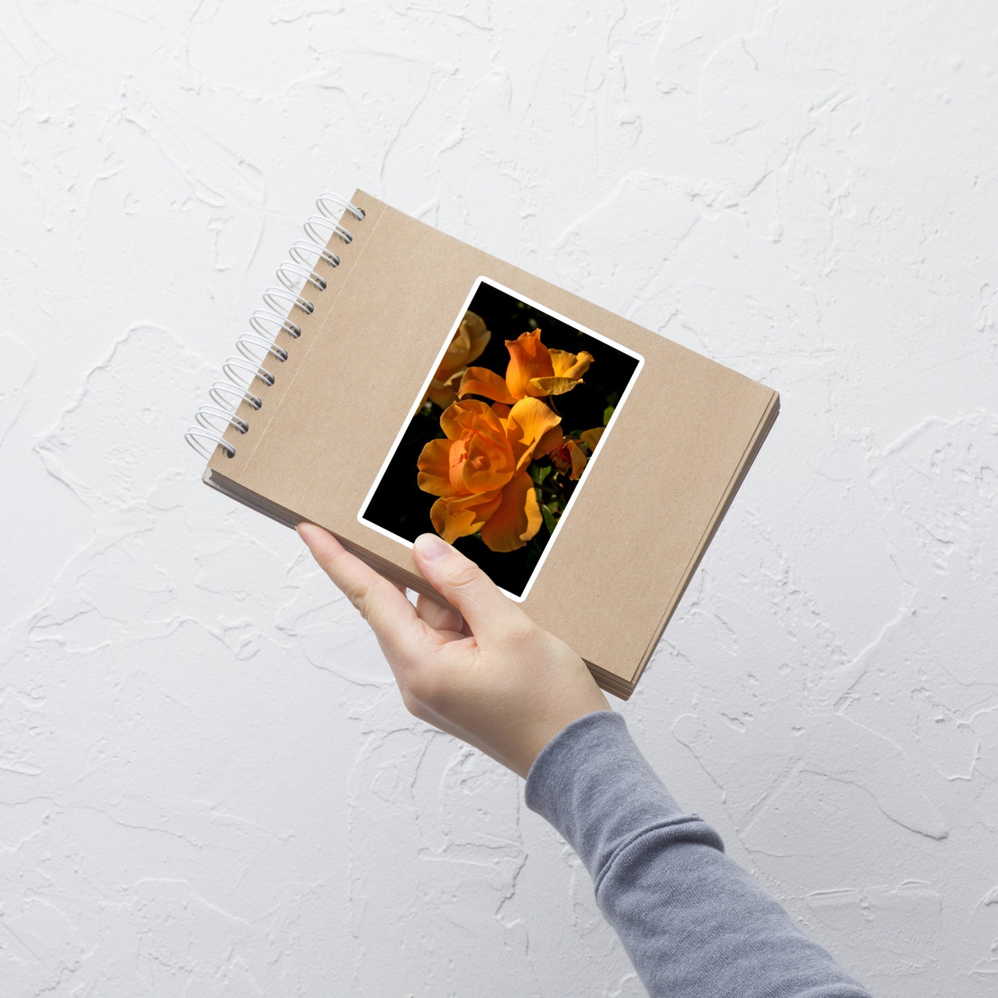 Lifestyle photo of a rectangular vinyl sticker with a high-contrast image of vivid orange roses against a dark background, placed on a small notepad.
