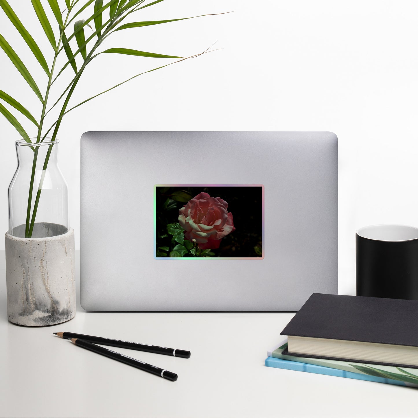 Lifestyle photo of a rectangular holographic sticker with a high-contrast image of a pink rose with dark green leaves, placed on a laptop with desk supplies nearby.