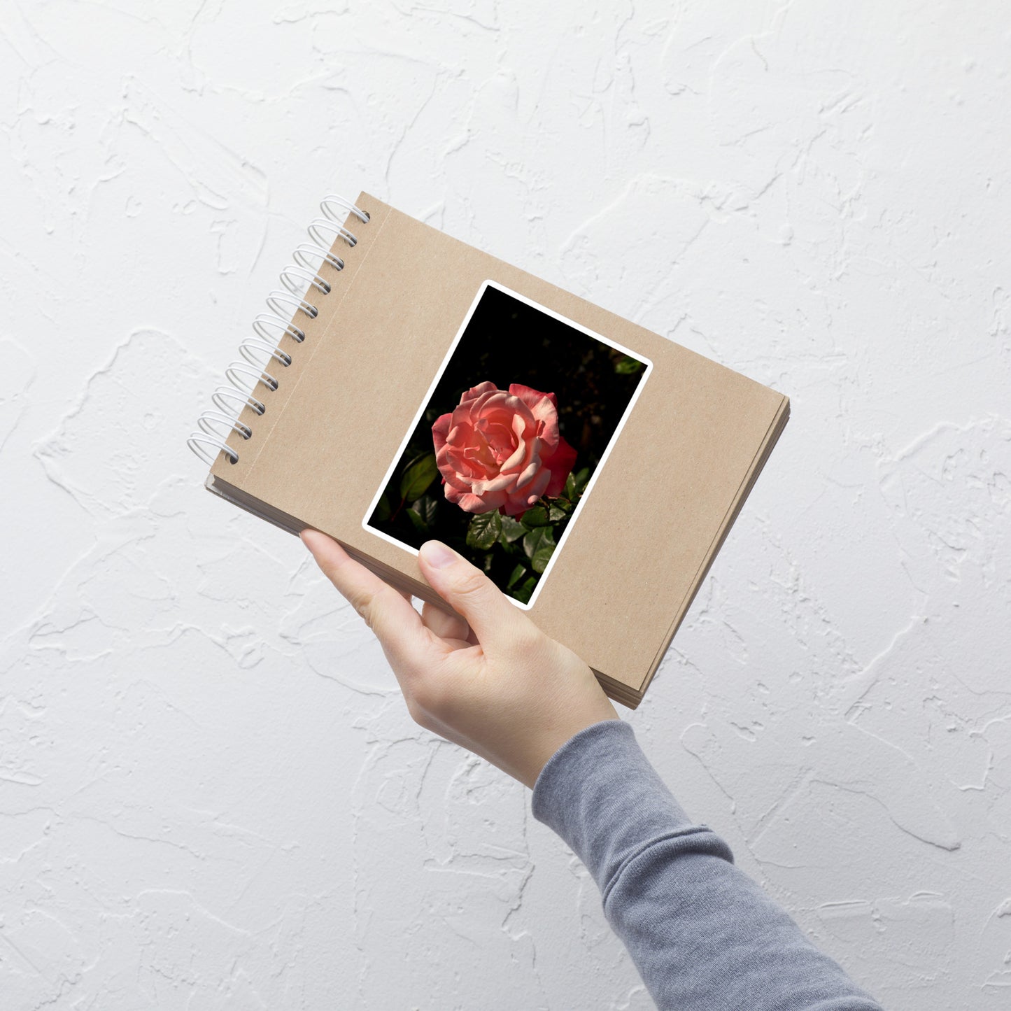 Lifestyle photo of a rectangular vinyl sticker with a high contrast image of a light pink rose bloom with dark green leaves, placed on a small notepad.