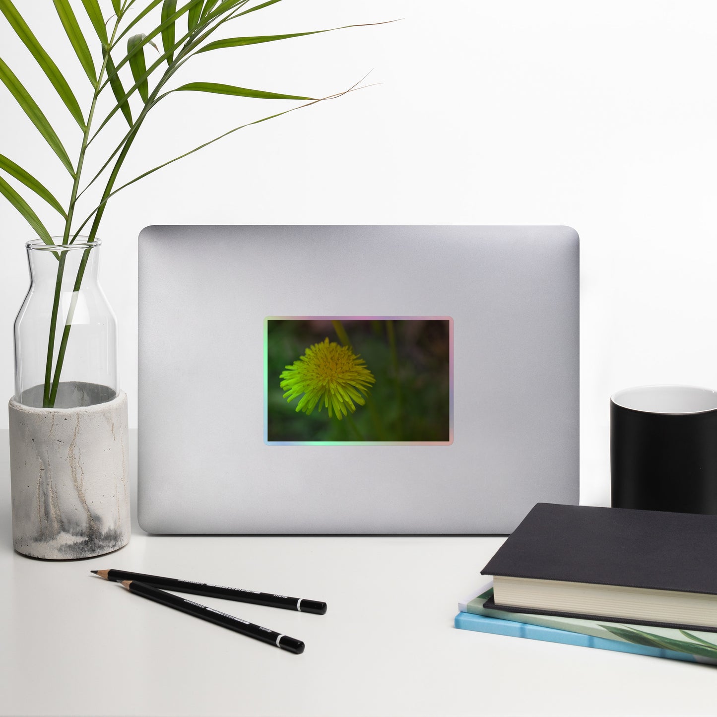 Lifestyle photo of a rectangular holographic sticker with a close-up image of a bright yellow dandelion bloom with a blurred light green background, placed on a laptop with desk supplies nearby.