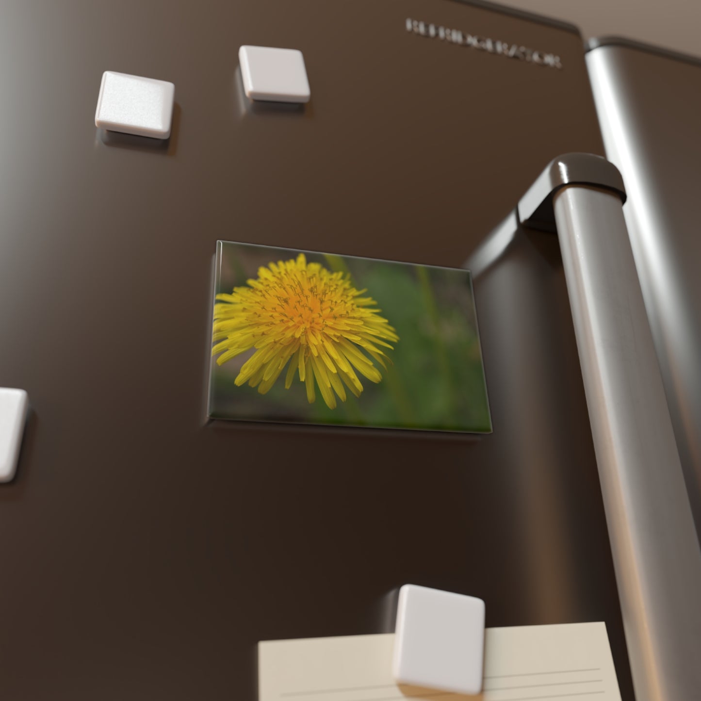 Lifestyle photo of a rectangular button magnet with a close-up image of a yellow dandelion flower, placed on a fridge.