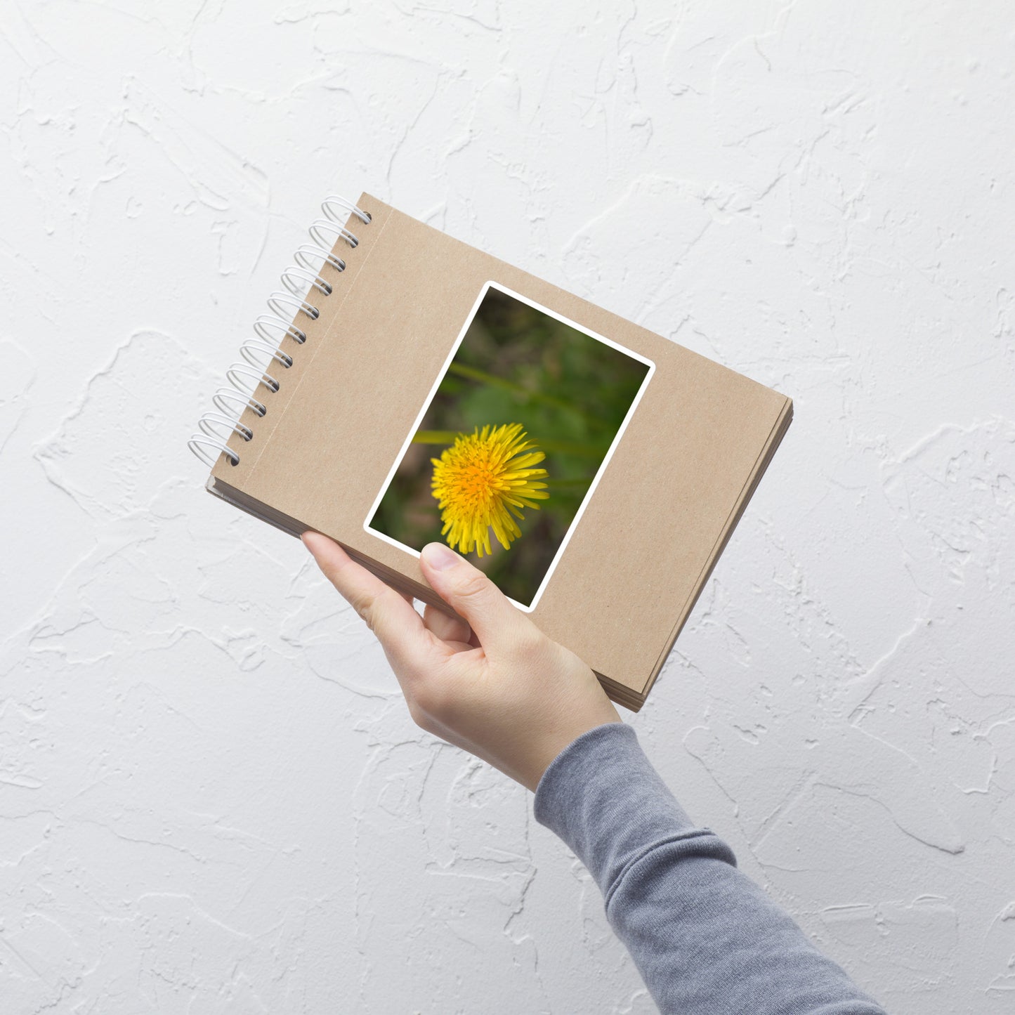 Lifestyle photo of a rectangular vinyl sticker with an image of a bright yellow dandelion bloom, placed on a small notepad.