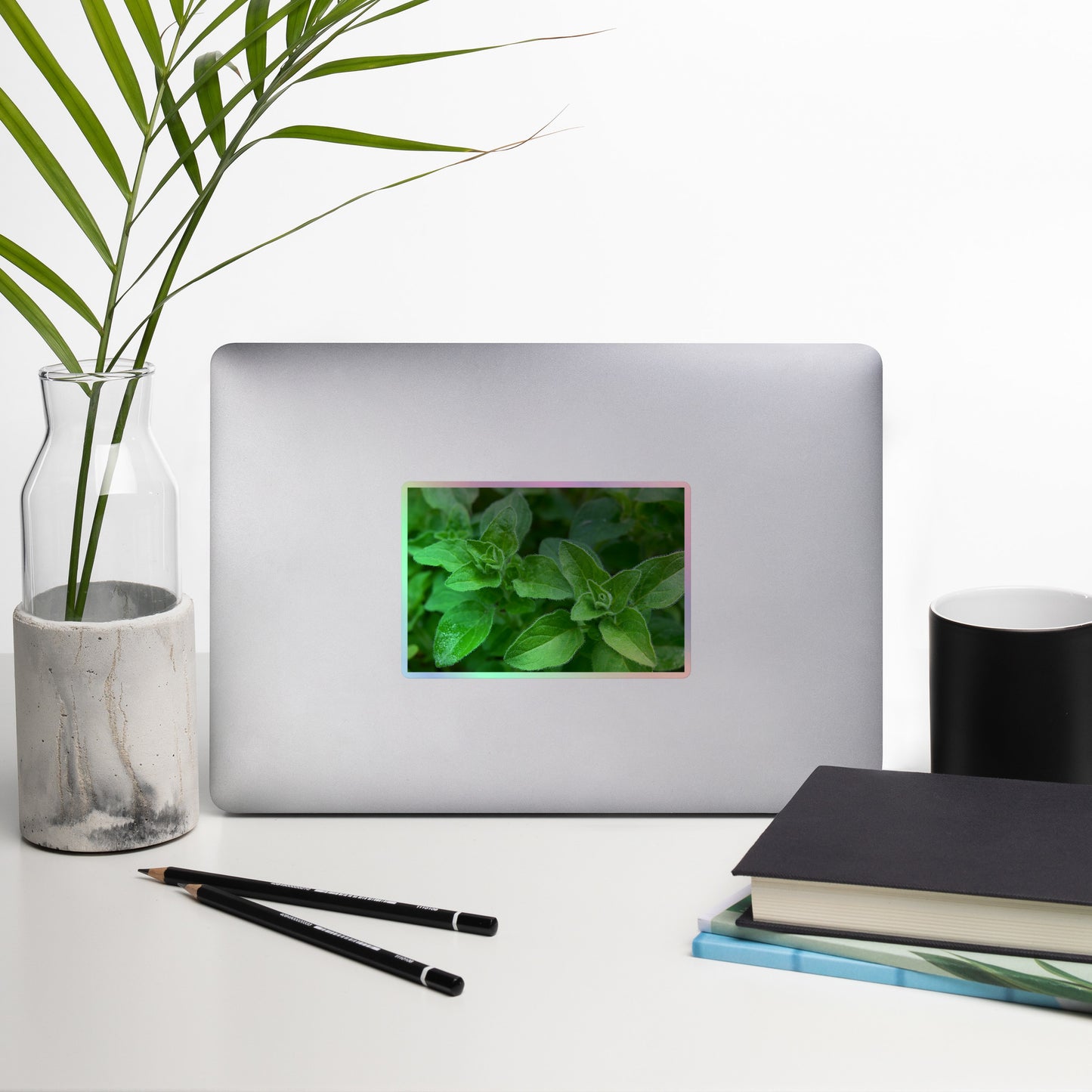 Lifestyle photo of a rectangular holographic sticker with a close-up image of vivid green oregano leaves, placed on a laptop with desk supplies nearby.