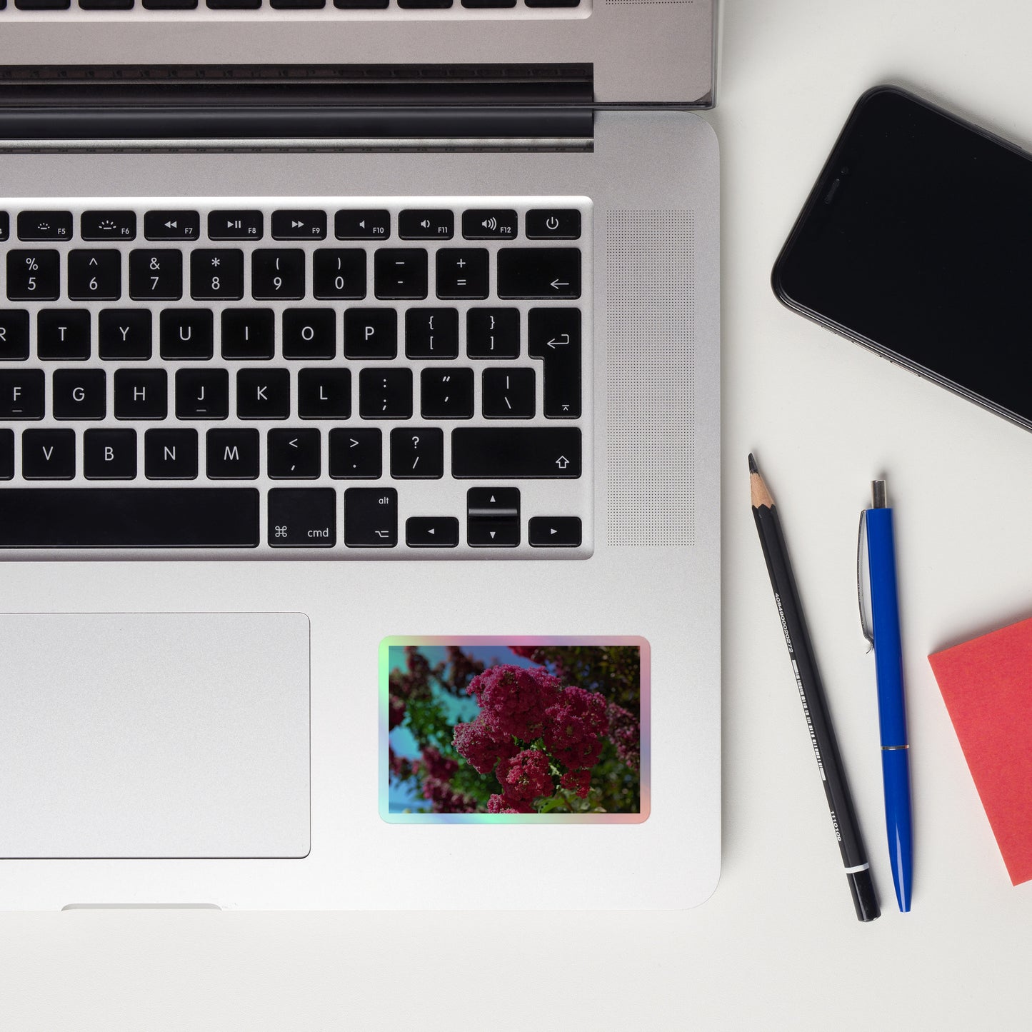 Lifestyle photo of a rectangular holographic sticker with an image of bright pink crape myrtle blooms with green leaves and blue sky in the background, placed on a laptop to the right of the mouse pad.