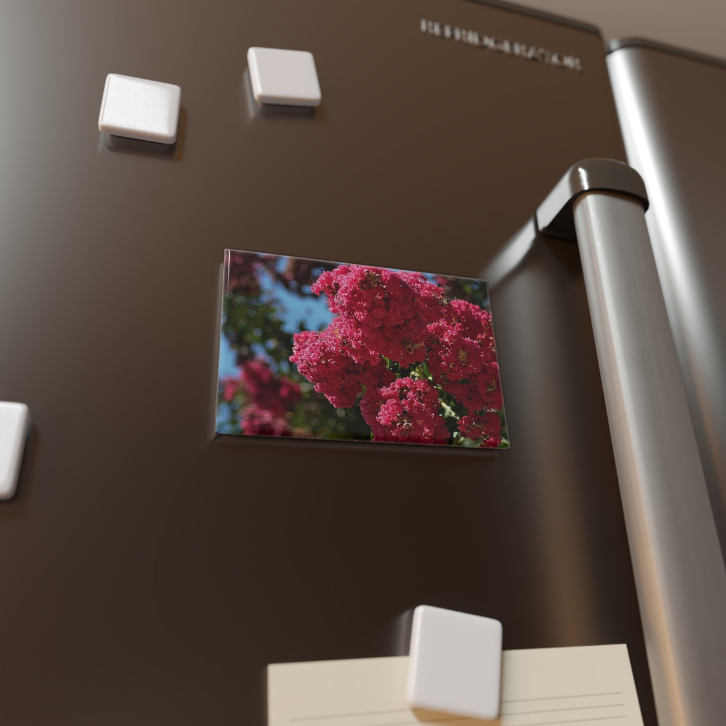 Lifestyle photo of a rectangular button magnet with an image of vibrant pink Crape Myrtle flowers in full bloom with green leaves and blue sky in the background, placed on a fridge.