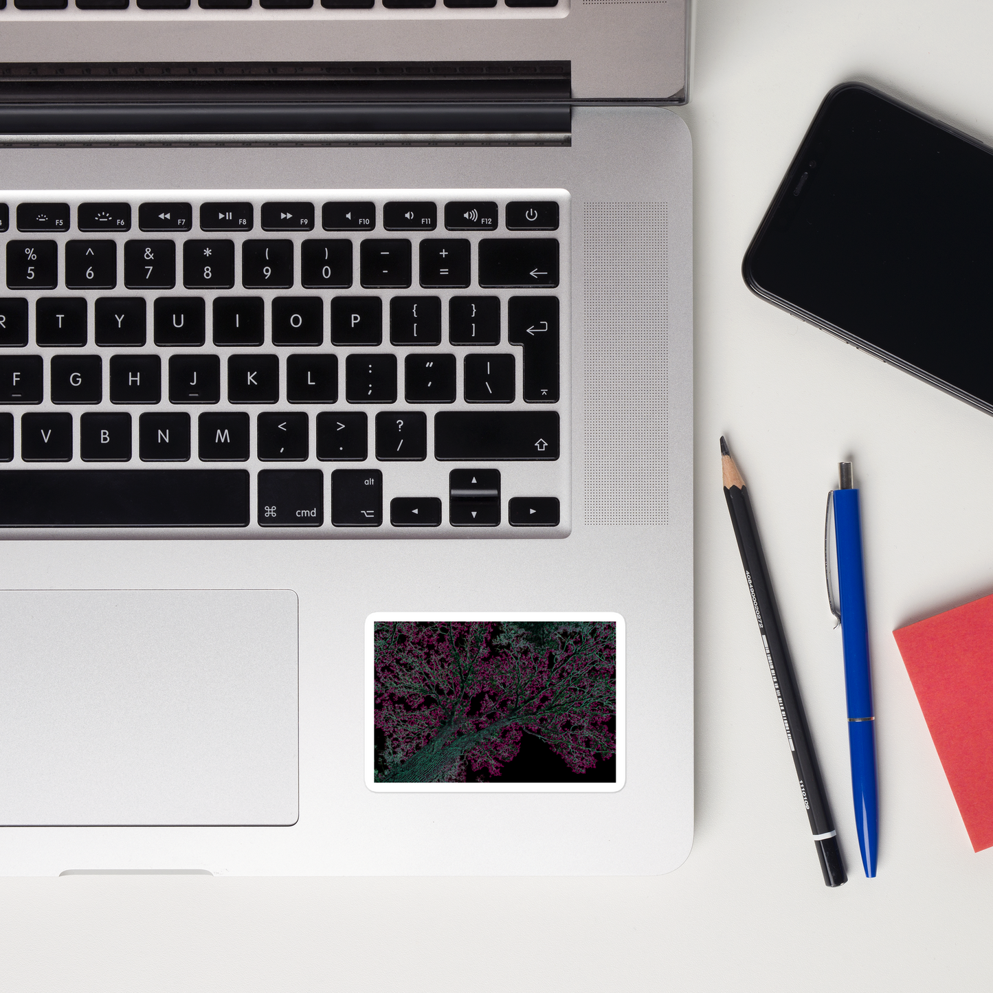 Lifestyle photo of a rectangular vinyl sticker with an image of a neon pink and turquoise tree design with lightning like features, placed on a laptop to the right of the mouse pad.