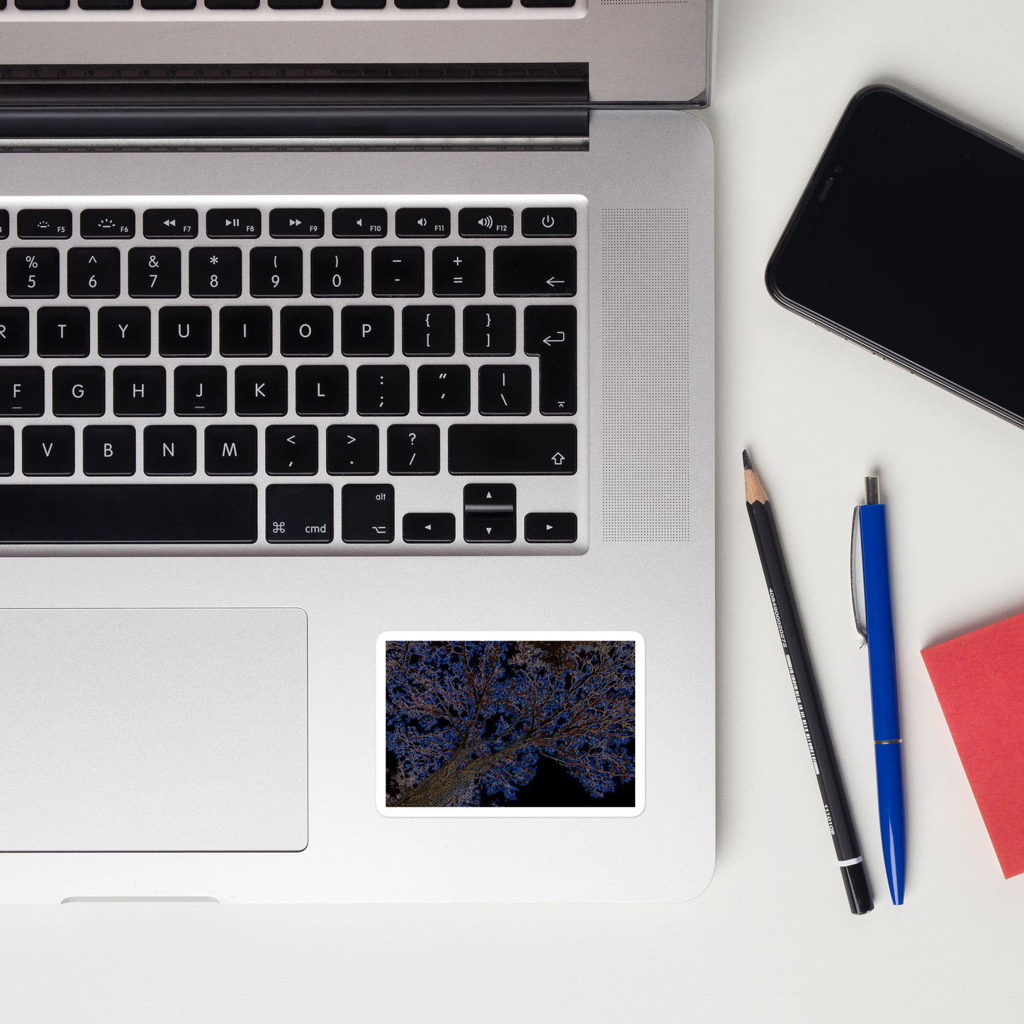 Lifestyle photo of a rectangular vinyl sticker with an image of a neon blue and yellow tree design with lightning like features, placed on a laptop to the right of the mouse pad.
