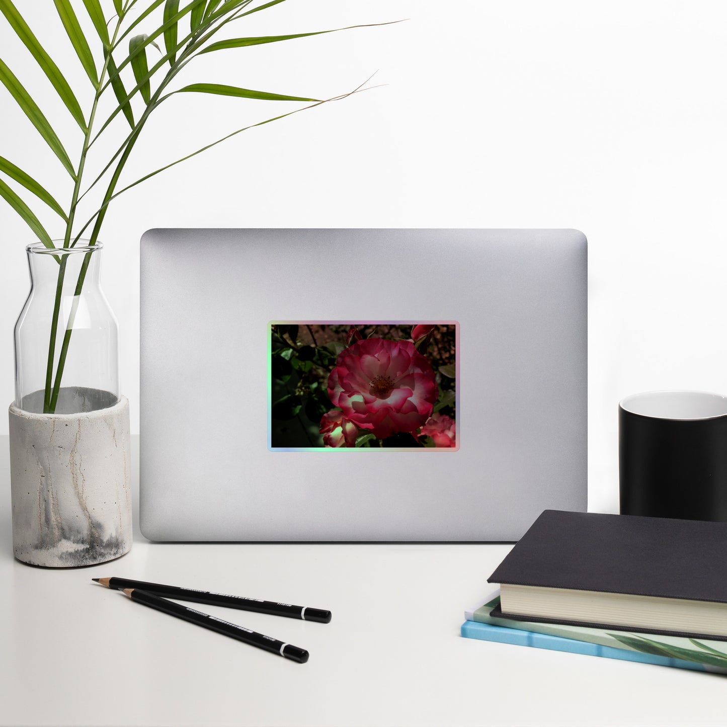 Lifestyle photo of a rectangular holographic sticker with an image of bright pink rose blooms with white centers, placed on a laptop with desk supplies nearby.