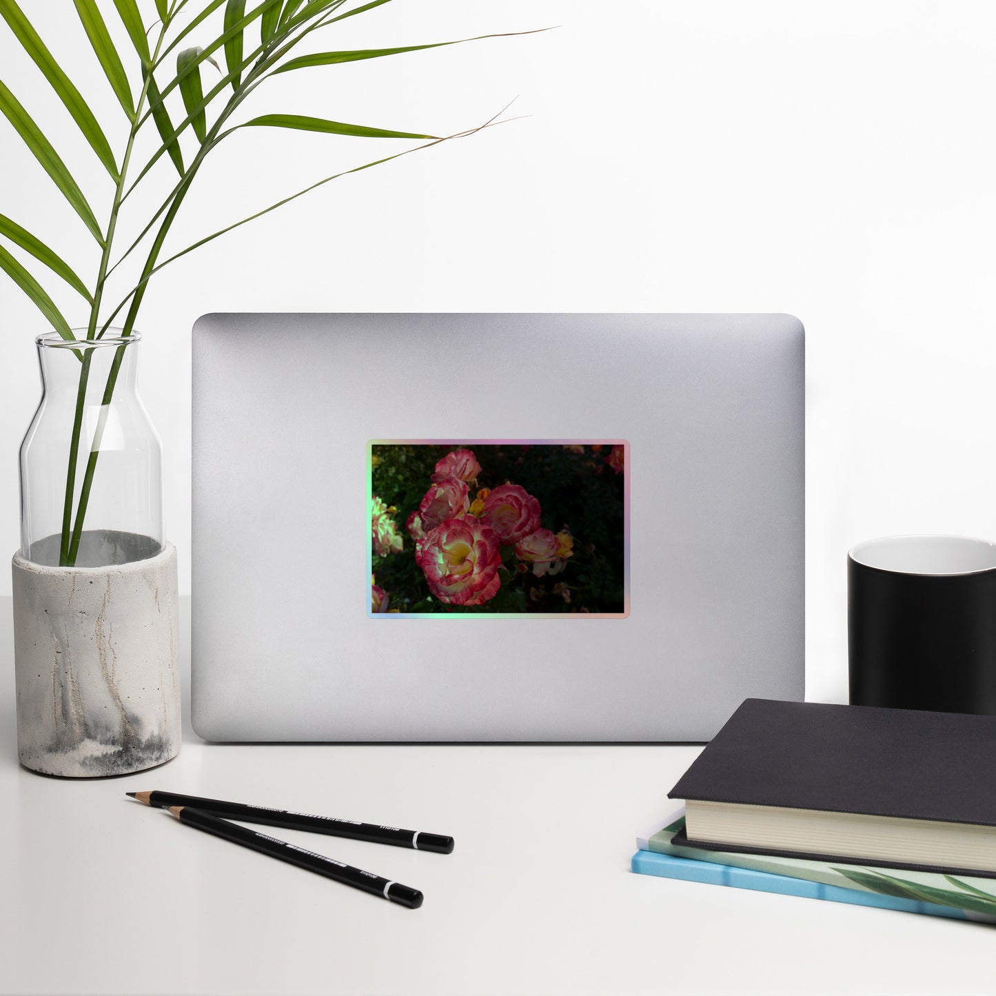 Lifestyle photo of a rectangular holographic sticker with an image of pink, yellow and white rose blooms with dark green leaves, placed on a laptop with desk supplies nearby.