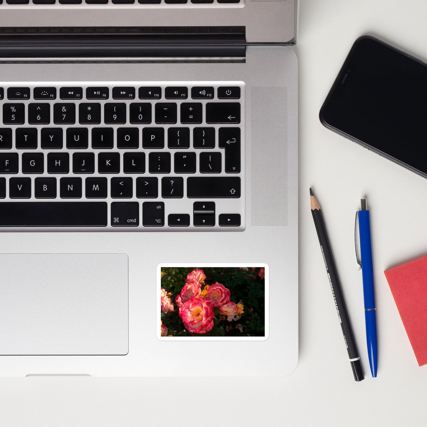 Lifestyle photo of a rectangular vinyl sticker with an image of a garden with pink, yellow, and white roses, placed on a laptop to the right of the mouse pad.
