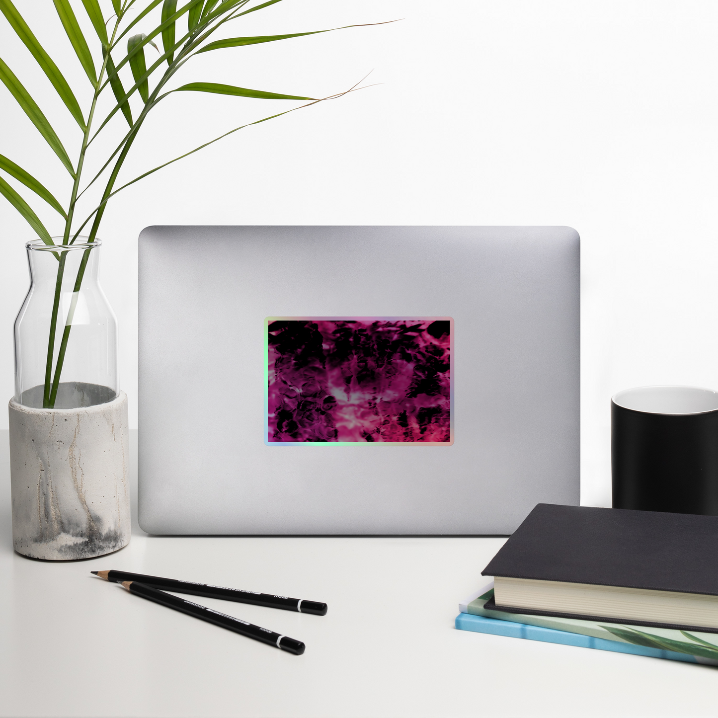 Lifestyle photo of a rectangular holographic sticker with an image of a vivid pink and black water reflection design, placed on a laptop with desk supplies nearby.