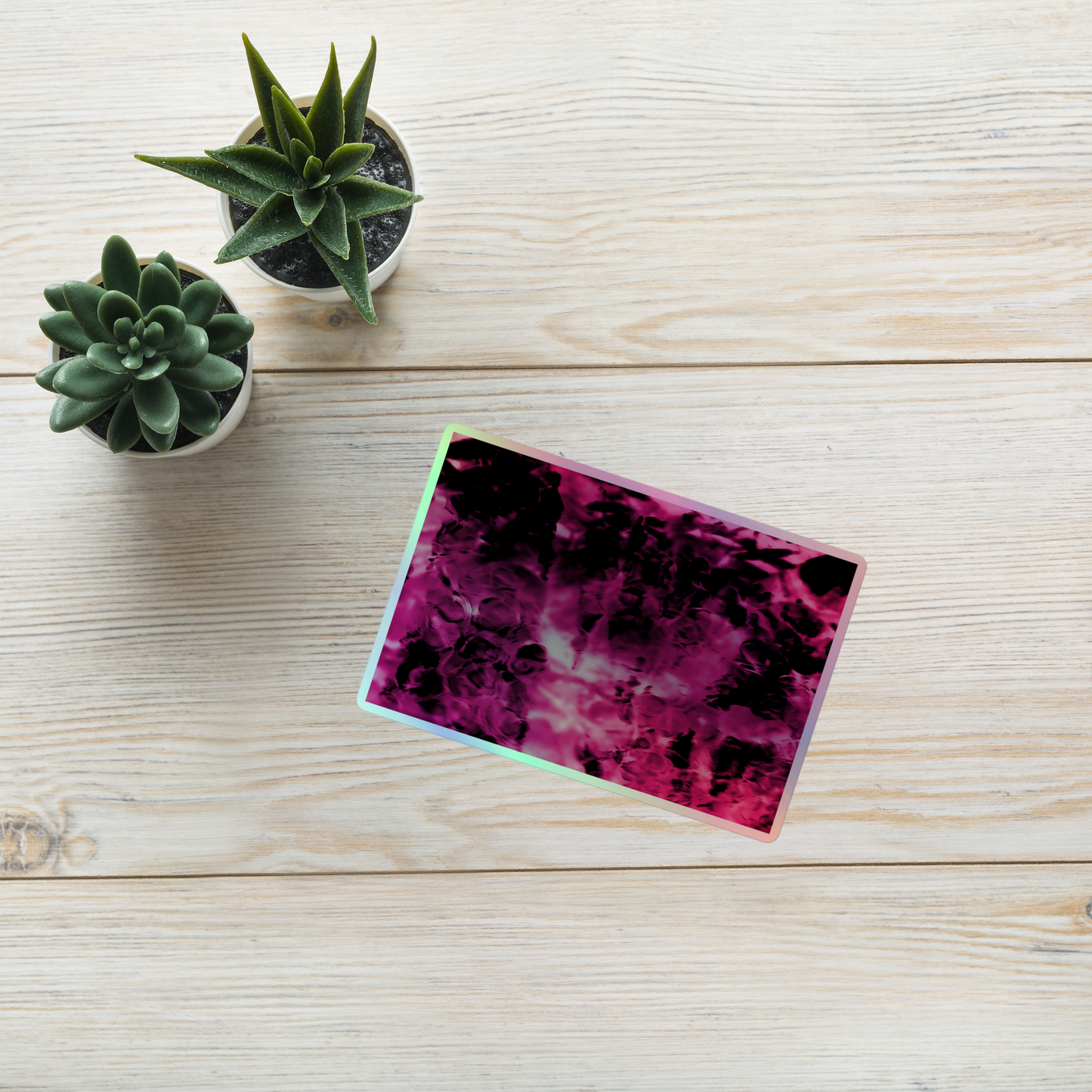 Lifestyle photo of a rectangular holographic sticker with an image of a vivid pink and black water reflection design, placed on a table with two small succulents.