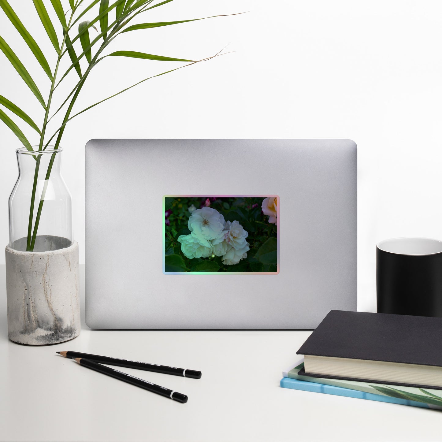 Lifestyle photo of a rectangular holographic sticker with an image of a cluster of white roses with dark green leaves, placed on a laptop with desk supplies nearby.