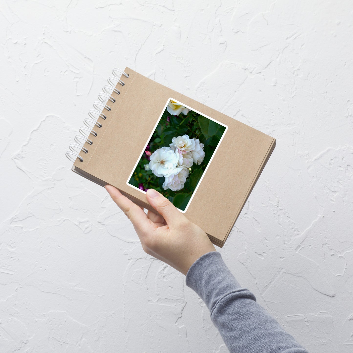 Lifestyle photo of a rectangular vinyl sticker with an image of white roses among dark green leaves, placed on a small notepad.