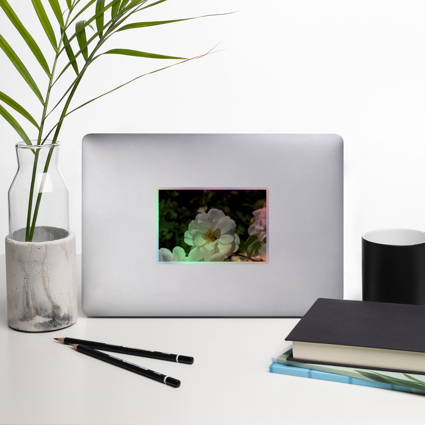 Lifestyle photo of a rectangular holographic sticker with an image of a white rose bloom with a honeybee on it, placed on a laptop with desk supplies nearby.