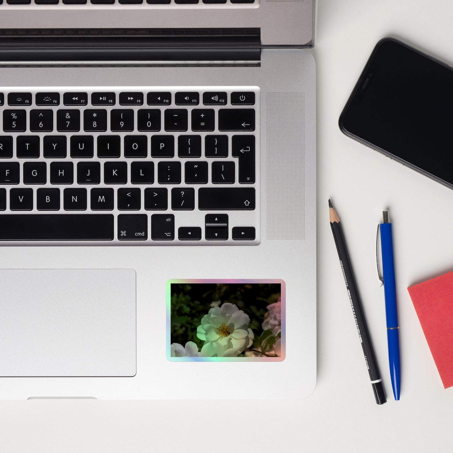 Lifestyle photo of a rectangular holographic sticker with an image of a white rose bloom with a honeybee on it, placed on a laptop to the right of the mouse pad.