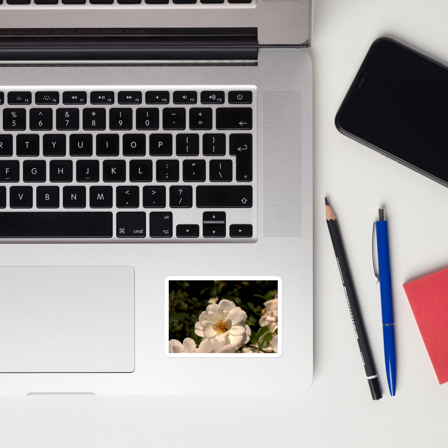Lifestyle photo of a rectangular vinyl sticker with an image of a white rose with a honeybee on it, placed on a laptop to the right of the mouse pad.