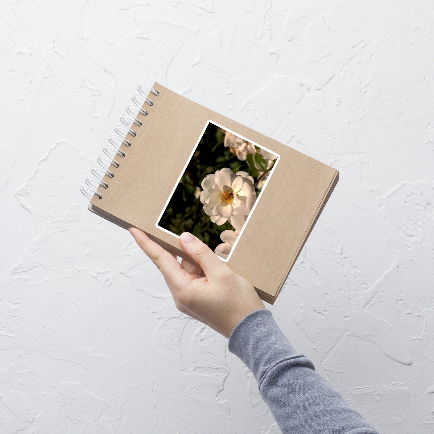 Lifestyle photo of a rectangular vinyl sticker with an image of a white rose with a honeybee on it, placed on a small notepad.