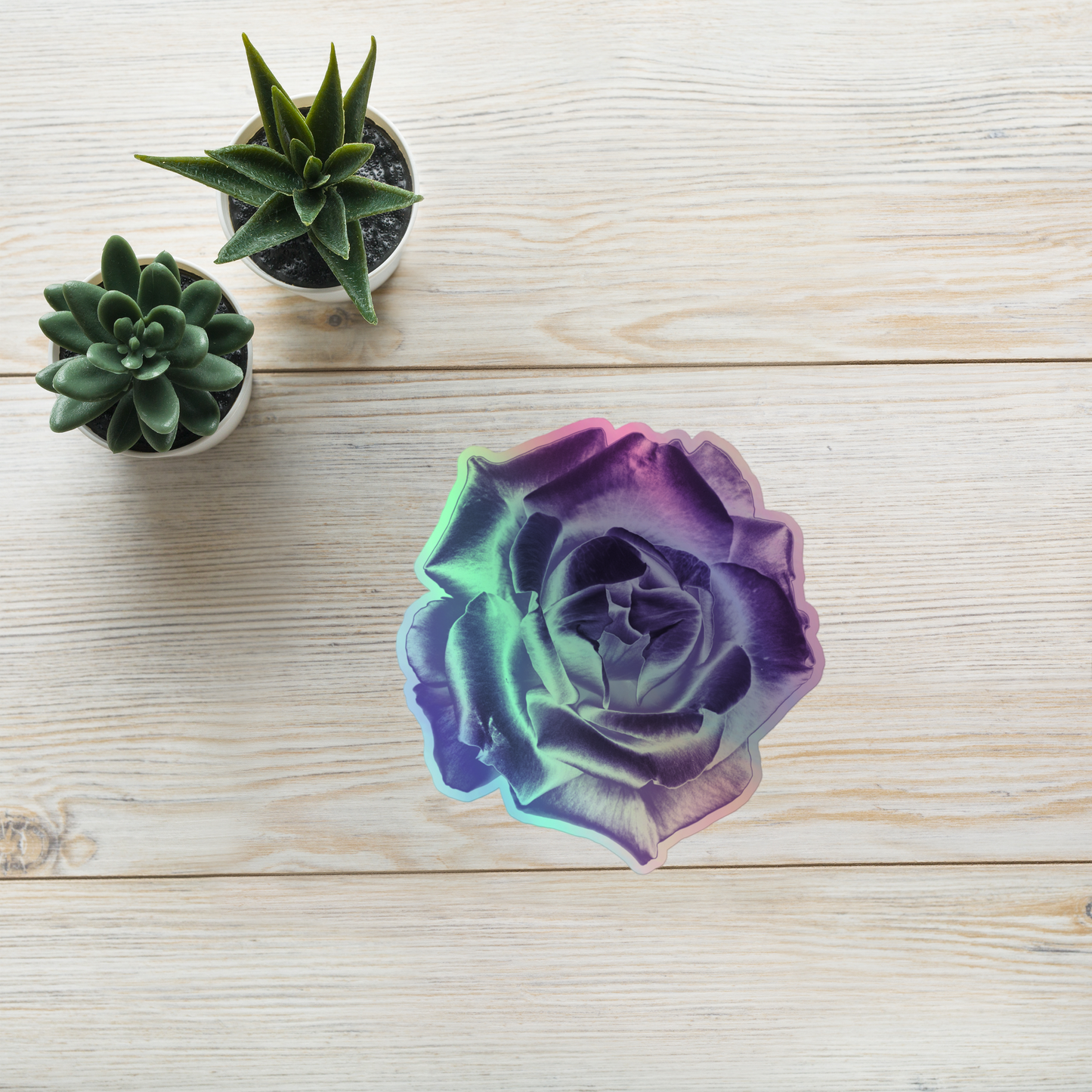 Lifestyle photo of a kiss-cut holographic sticker with an image of a reflective purple rose design, placed on a table with two small succulents.