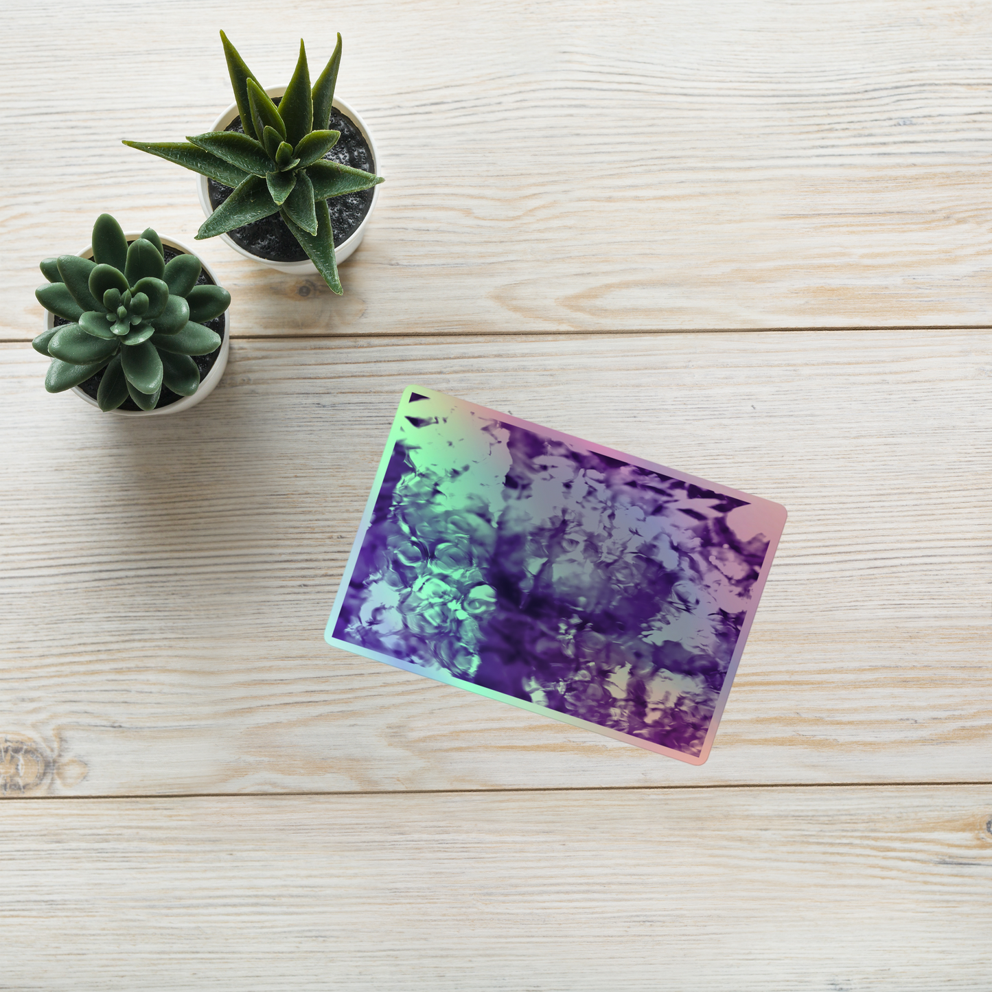 Lifestyle photo of a rectangular holographic sticker with an image of a vivid purple water reflection design, placed on a table with two small succulents.