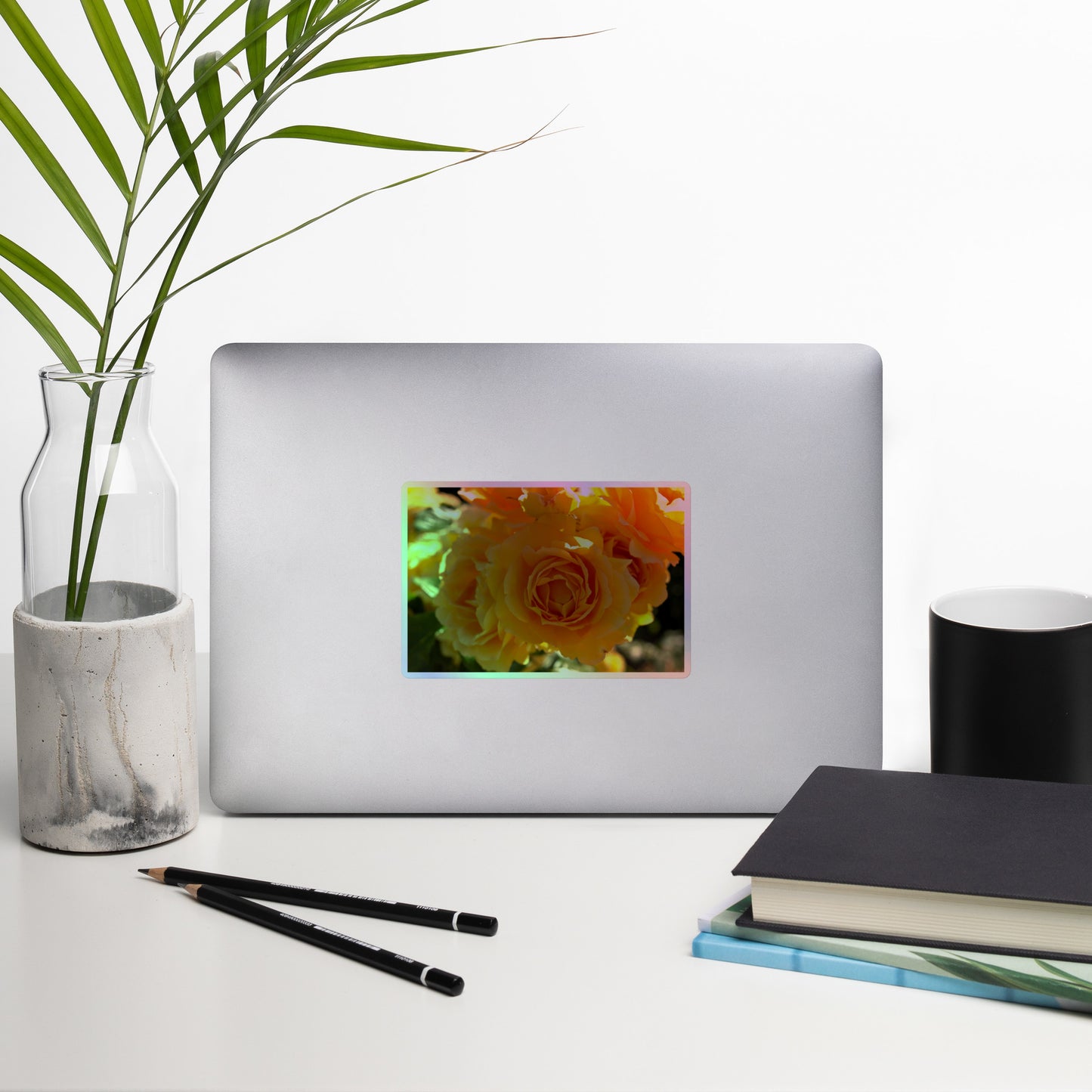 Lifestyle photo of a rectangular holographic sticker with an image of bright orange rose blooms in the sunlight, placed on a laptop with desk supplies nearby.