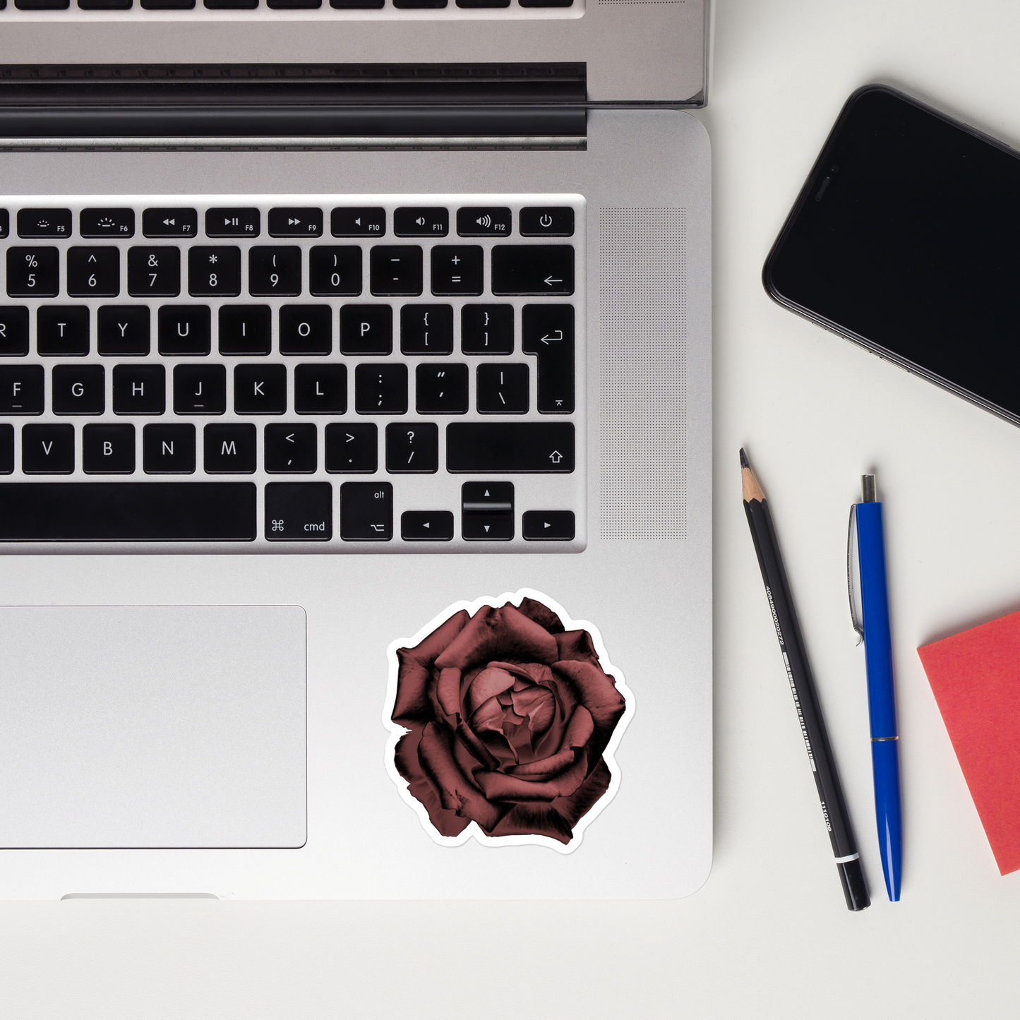 Lifestyle photo of a kiss-cut vinyl sticker with an image of a red charcoal drawing style rose, placed on a laptop to the right of the mouse pad.