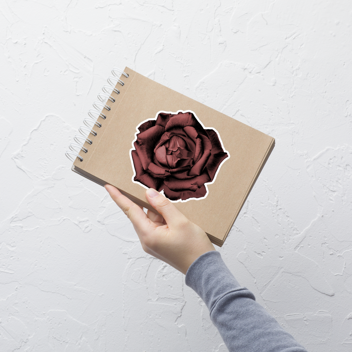Lifestyle photo of a kiss-cut vinyl sticker with an image of a red charcoal drawing style rose, placed on a small notepad.