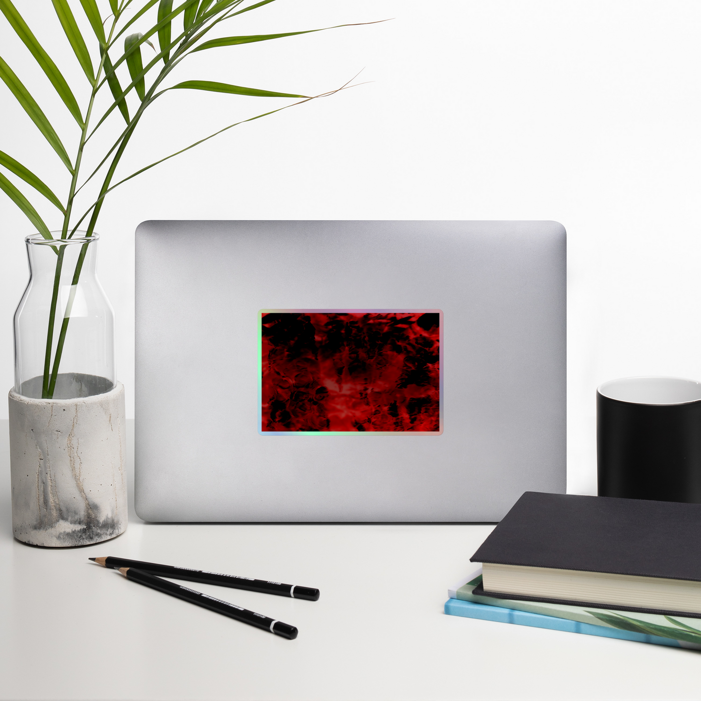 Lifestyle photo of a rectangular holographic sticker with an image of a vivid red and black water reflection design, placed on a laptop with desk supplies nearby.