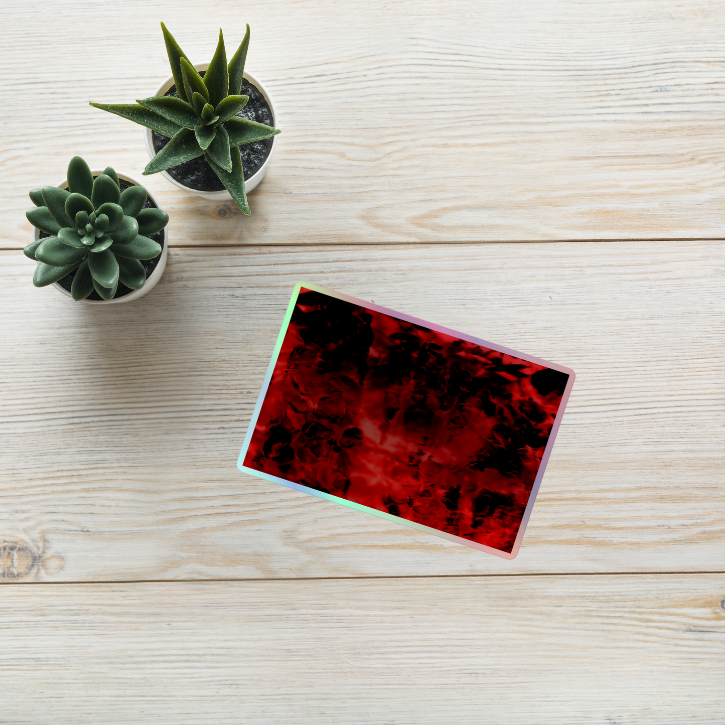 Lifestyle photo of a rectangular holographic sticker with an image of a vivid red and black water reflection design, placed on a table with two small succulents.