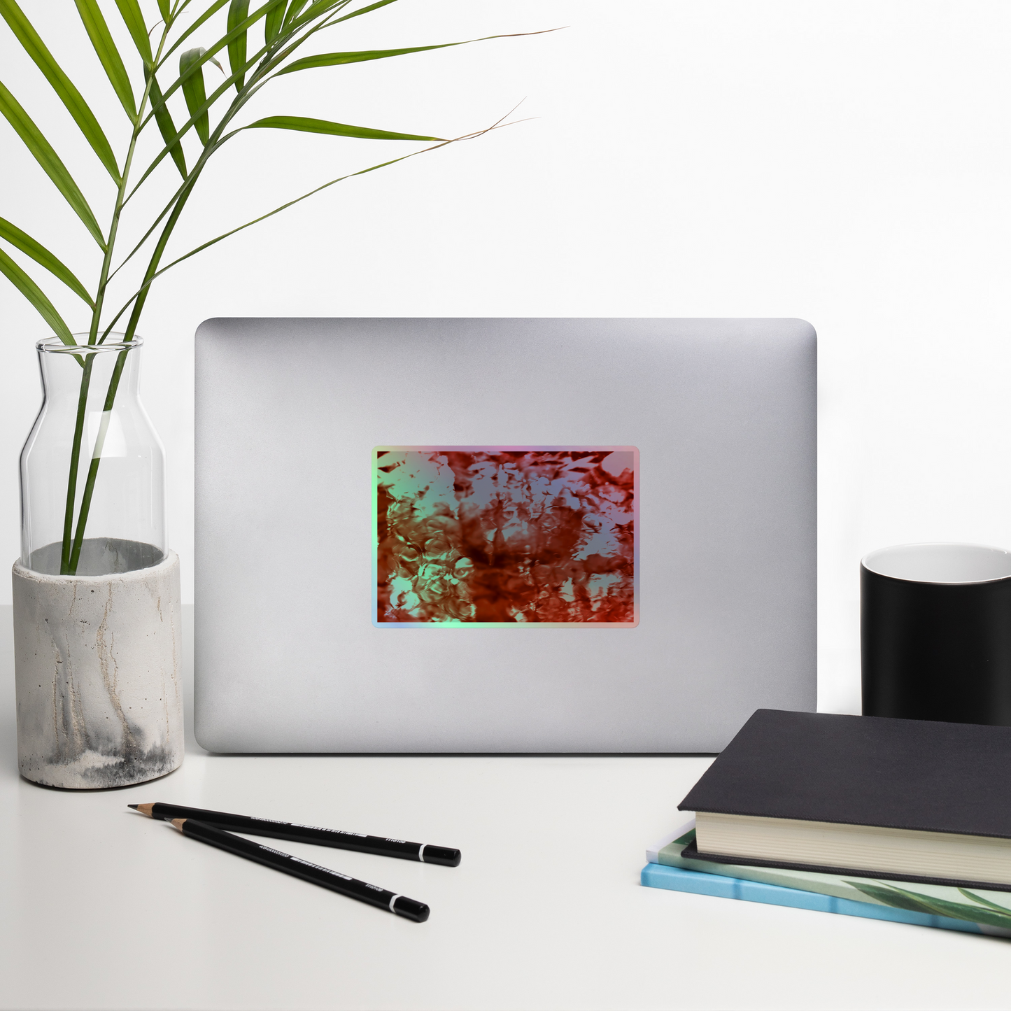Lifestyle photo of a rectangular holographic sticker with an image of a vivid red water reflection design, placed on a laptop with desk supplies nearby.