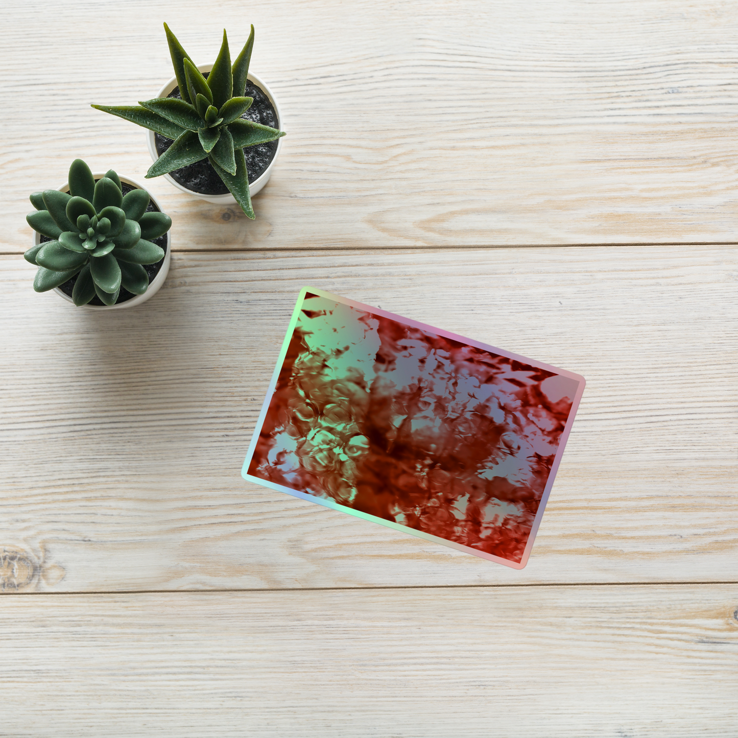 Lifestyle photo of a rectangular holographic sticker with an image of a vivid red water reflection design, placed on a table with two small succulents.