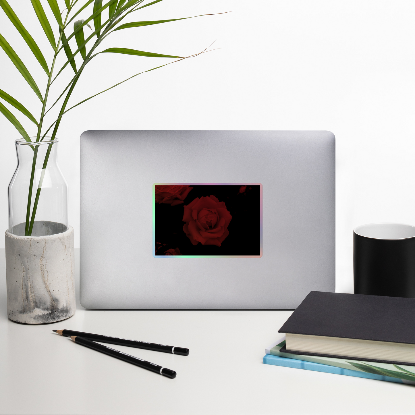 Lifestyle photo of a rectangular holographic sticker with an image of a red vivid rose design, placed on a laptop with desk supplies nearby.