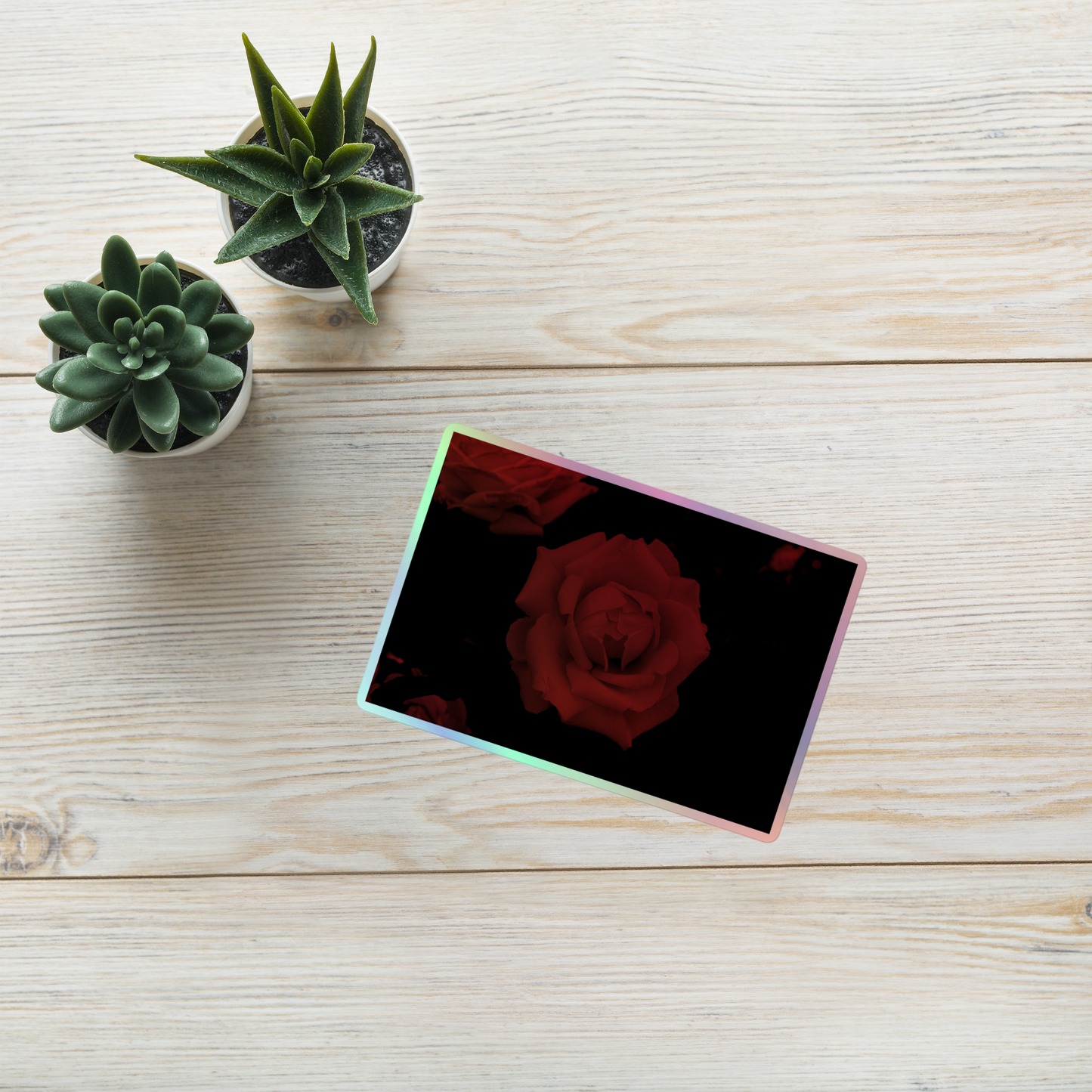 Lifestyle photo of a rectangular holographic sticker with an image of a red vivid rose design, placed on a table with two small succulents.