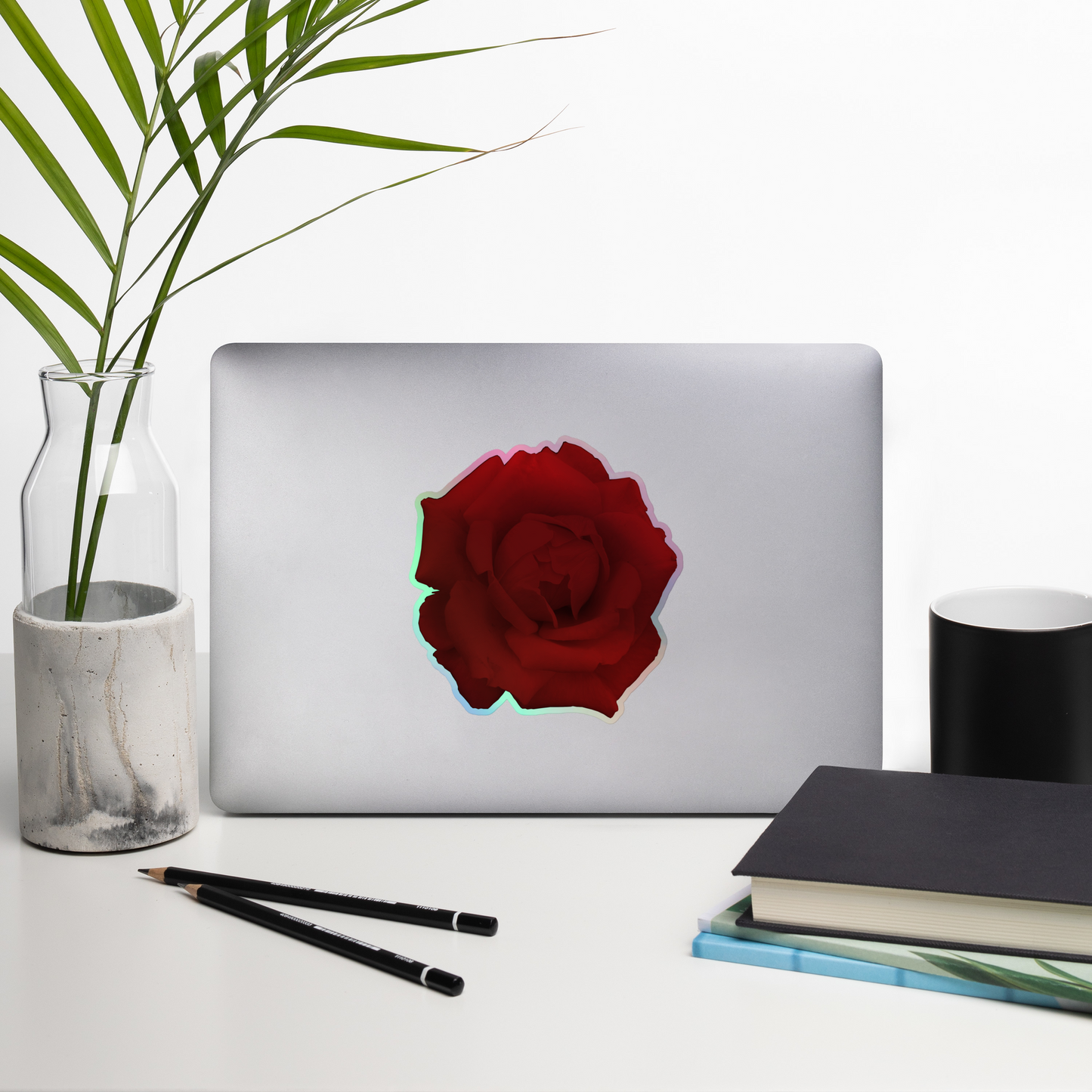 Lifestyle photo of a kiss-cut holographic sticker with an image of a vivid red rose design, placed on a laptop with desk supplies nearby.
