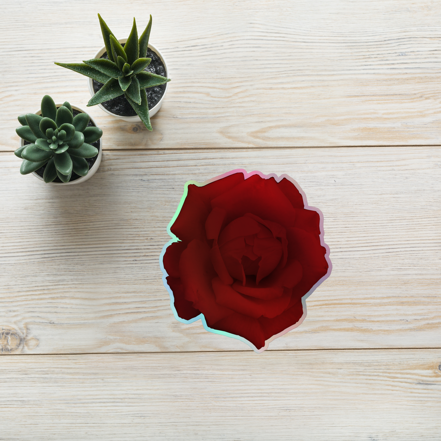 Lifestyle photo of a kiss-cut holographic sticker with an image of a vivid red rose design, placed on a table with two small succulents.
