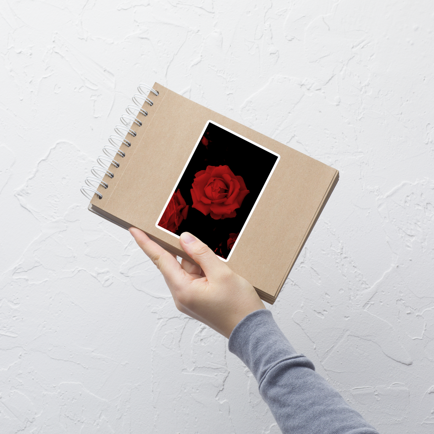Lifestyle photo of a rectangular vinyl sticker with an image of a vivid red rose design, placed on a small notepad.