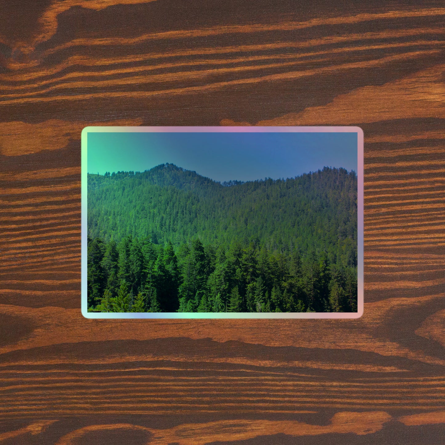 Lifestyle photo of a rectangular holographic sticker with a landscape image of blue sky, mountains, and green redwood trees, placed on a brown wooden table surface.
