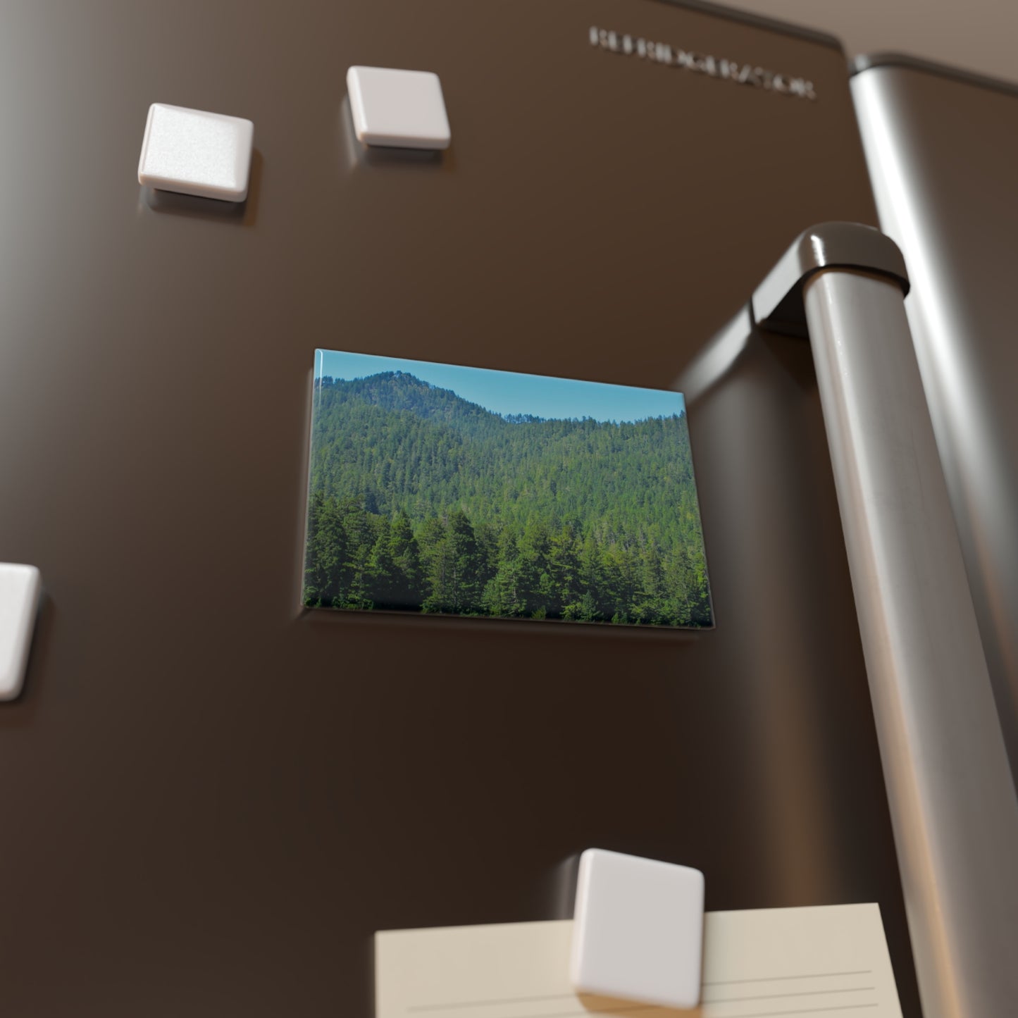 Lifestyle photo of a rectangular button magnet with an image of a dense green redwood forest on a mountain range, placed on a fridge.
