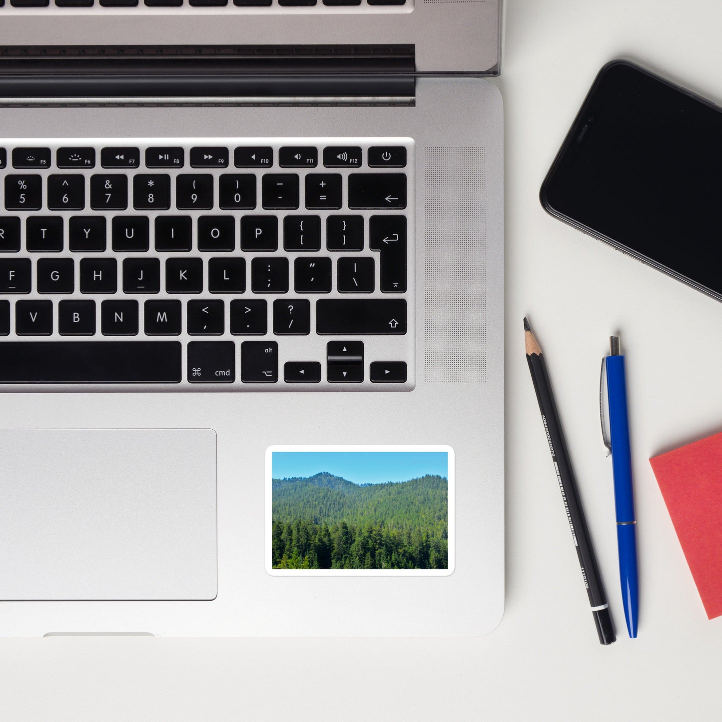 Lifestyle photo of a rectangular vinyl sticker with an image of mountain landscape with redwood trees and blue sky, placed on a laptop to the right of the mouse pad.